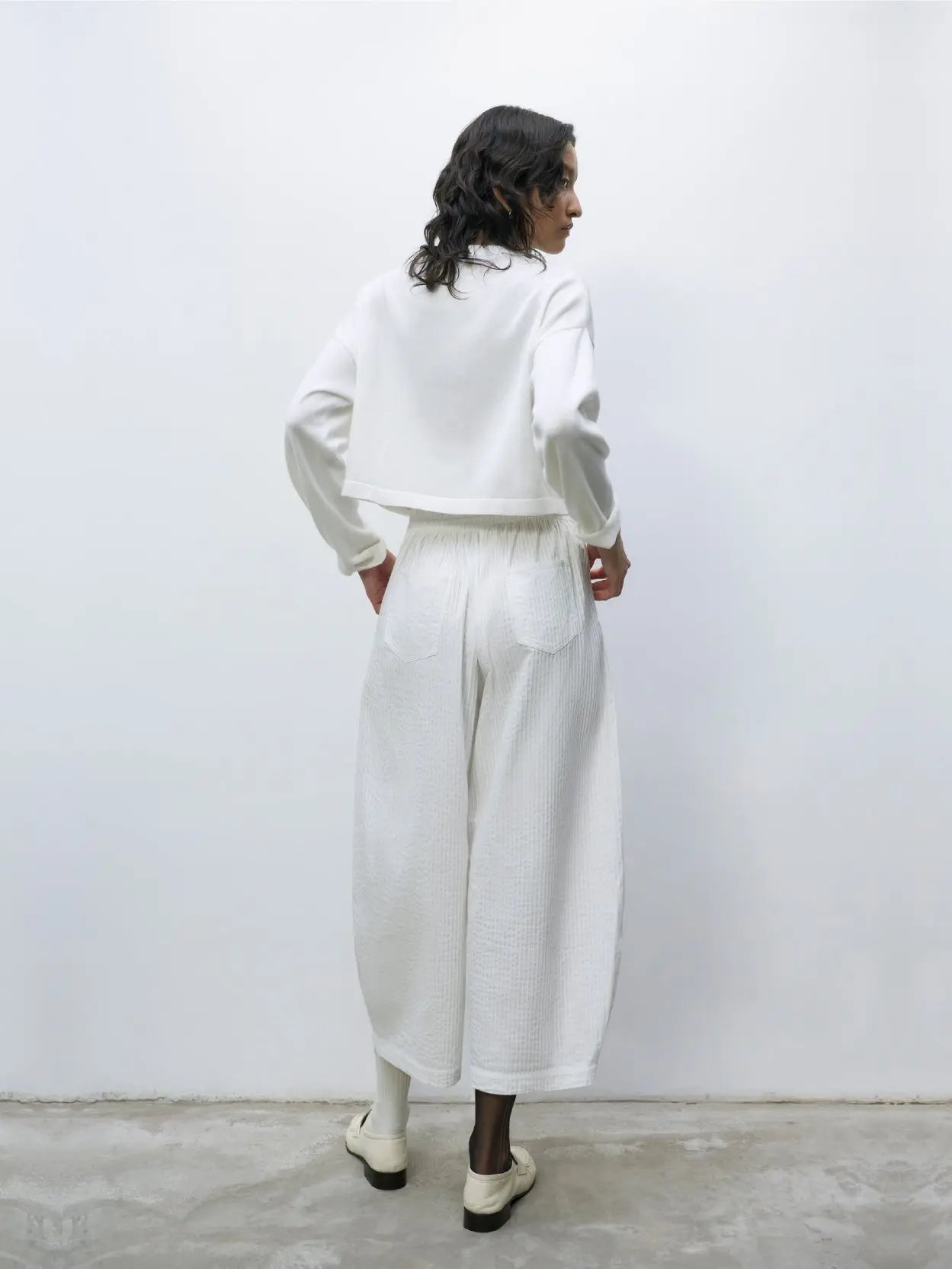 A white, long-sleeved, cropped cardigan with buttons down the front from Cordera. The Cotton Cropped Cardigan White has a simple, minimalist design with a round neckline and ribbed cuffs at the sleeves. The fabric appears soft and lightweight against a plain white background.