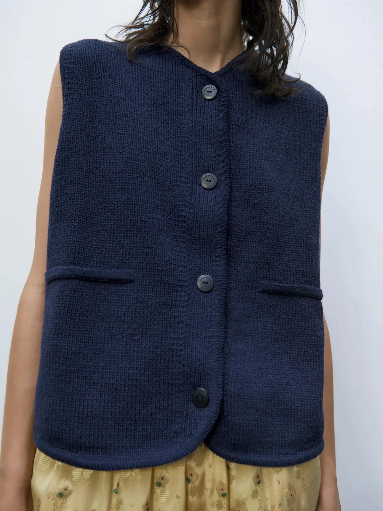 A dark blue, sleeveless vest with a simple design is available at Bassalstore in Barcelona. The vest features a button-down front with four buttons and two small pockets at the lower front. The fabric has a textured appearance. The back is not visible. The label inside the collar reads "Cotton Waistcoat Navy" by Cordera.