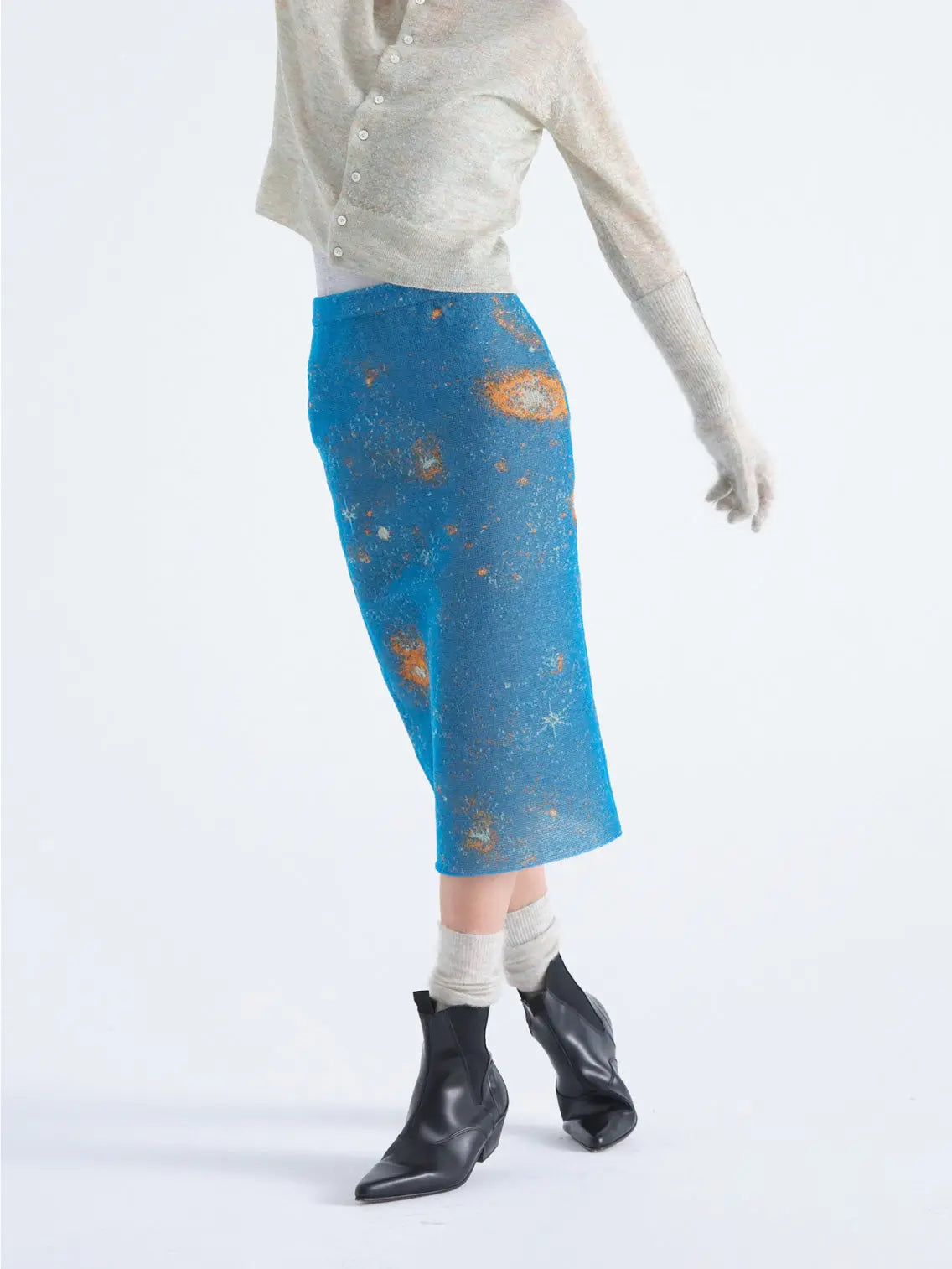 A knee-length blue skirt with an abstract design featuring orange patches and white star-like patterns, creating a visually dynamic galaxy-inspired look. The texture appears fuzzy, giving it a cozy and unique feel. The Galaxy Skirt Blue by Bielo is available at BassalStore in Barcelona for those seeking distinctive style.