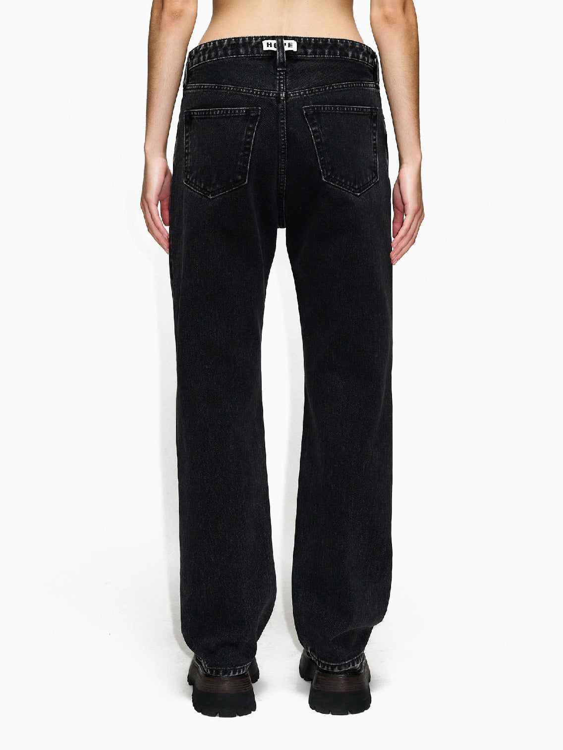 A person wearing Hope's Easy Jeans Chipped Black, high-waisted and loose-fitting, made from sustainable denim and paired with black shoes, stands with arms relaxed by their sides against a white background. These jeans, crafted from organic cotton, feature front pockets, belt loops, and a button/zipper closure. The person's torso is not shown.