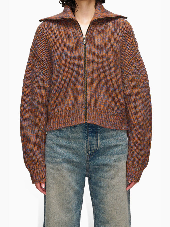 The Yummy Cardigan Orange by Hope is a cropped, brown, and orange knit sweater featuring a high collar and central zipper. This bomber jacket-inspired cardigan has long sleeves, a slightly relaxed fit, and is crafted from heavy wool yarn for warmth.