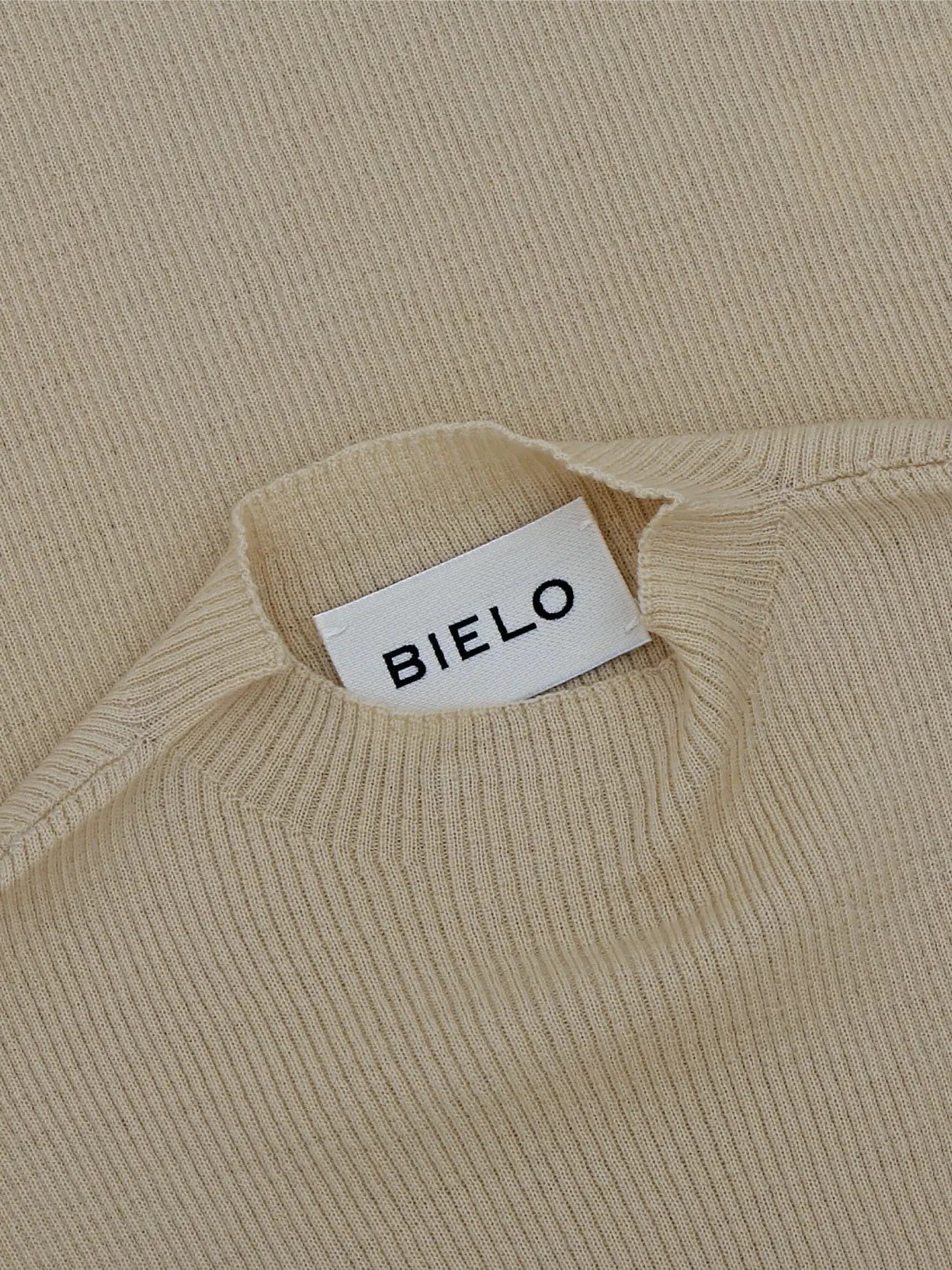 A sleeveless, ribbed-knit beige top with a high, rounded neckline. The Matte Top Cream from Bielo extends to a mid-length and has a simplistic, minimalistic design. The background is plain white.