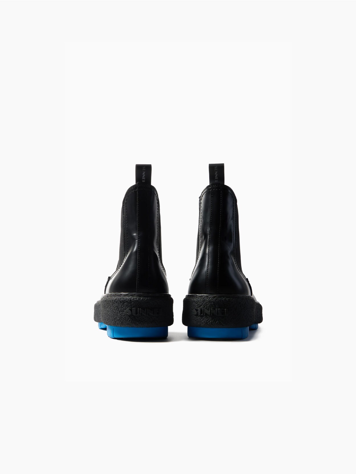 The Cemento Chelsea Boot by Sunnei is a black leather ankle boot with a chunky textured sole. It features black detailing and a vibrant blue tread, along with elastic side panels and pull tabs for easy wear, all set against a plain white background.