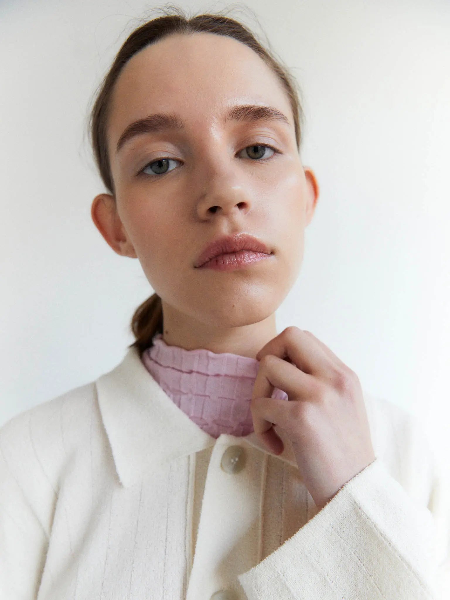 A cream-colored, long-sleeve button-up cardigan with a ribbed texture. It features a collar, front buttons, and a single large pocket on the left side. The label inside the neckline reads "Rus." Laid flat against a plain white background, this stylish piece is available at Bassalstore in Barcelona. This is the Tobira Cardigan Chalk by Rus.