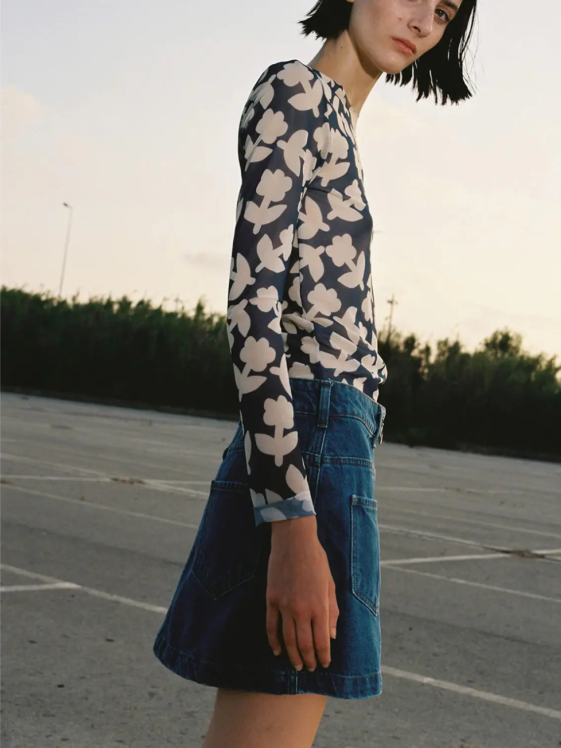 A person is standing outdoors wearing a long-sleeve top with a white floral pattern on a dark background and a Denim Mini Skirt from Mundaka, featuring a front zipper and large pockets. The top has a fitted design, and the skirt ends above the knees.
