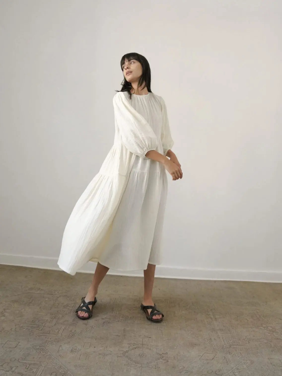 A person with long dark hair is standing in a minimalist room with white walls in Barcelona. They are wearing the Nella Dress by Zii Ropa with black strappy sandals. Their expression is neutral as they look directly at the camera.