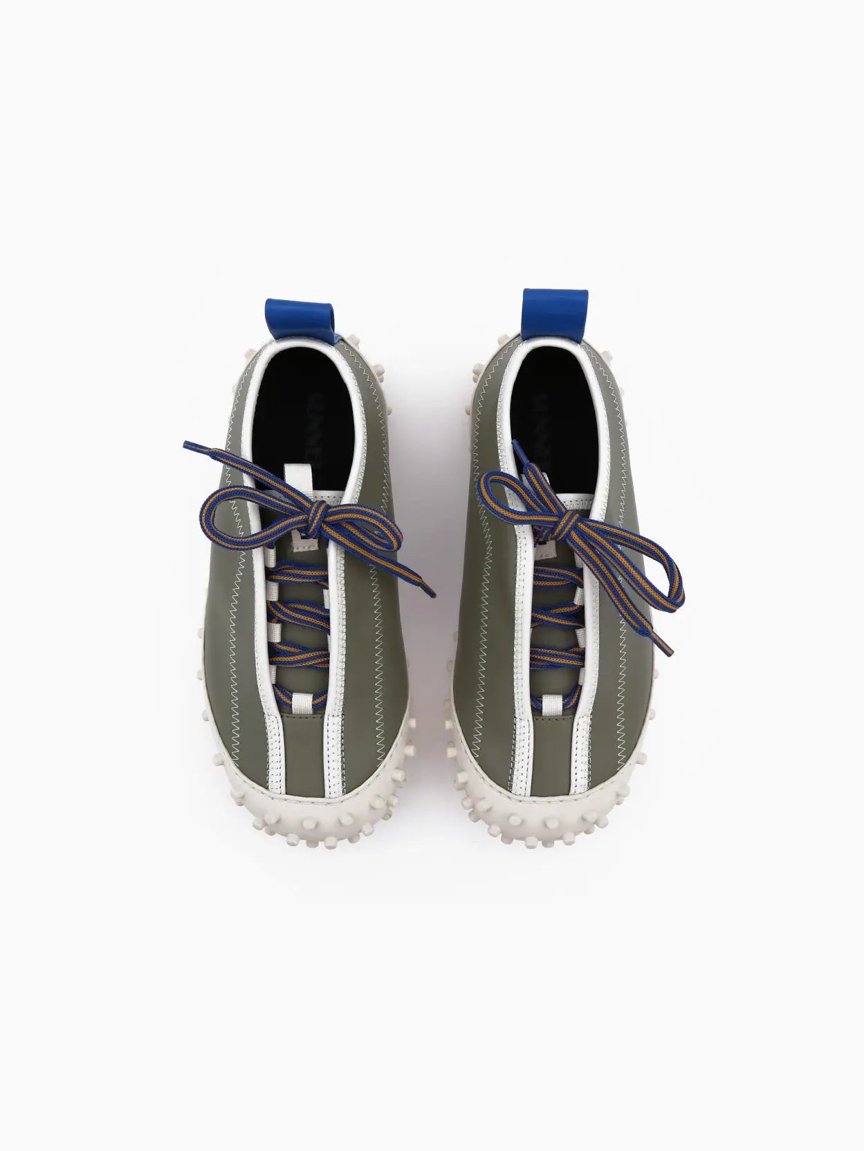 A single, stylish slip-on shoe with a gray and white color scheme and a blue heel tab. Available at Bassalstore, the Sunnei 1000 Chiodi Trainers Military Green features a unique, textured white sole with numerous round, prominent grips for enhanced traction. The lace has a colorful pattern and is tied neatly on top.