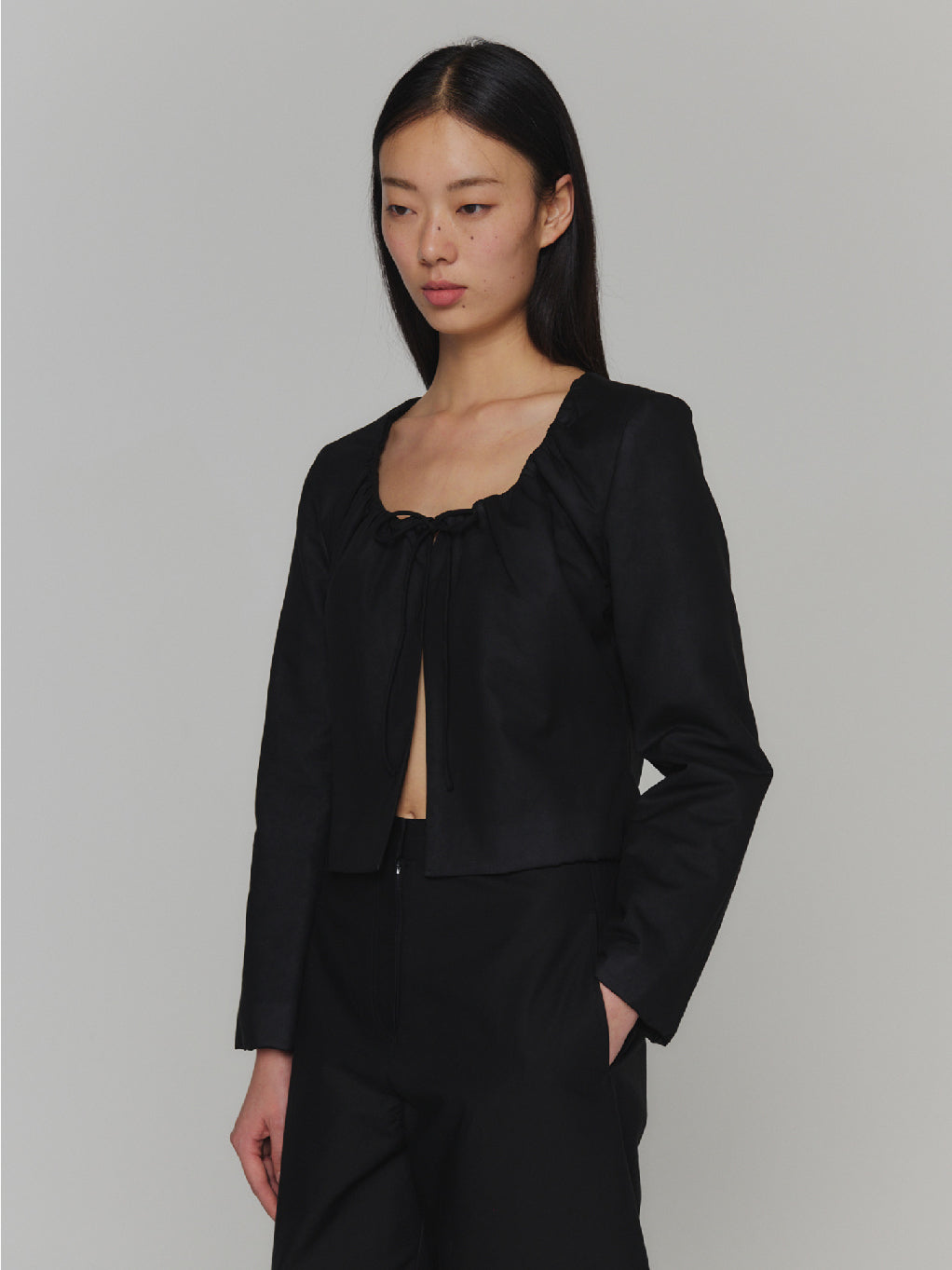 A "Sheer Padded Cropped Jacket Black" by Amomento displayed on a white background. The jacket, with its black color and long sleeves, boasts an adjustable square neckline, natural shirring near the chest area, and a subtle bow detail at the front. Its minimalistic design is both elegant and contemporary.