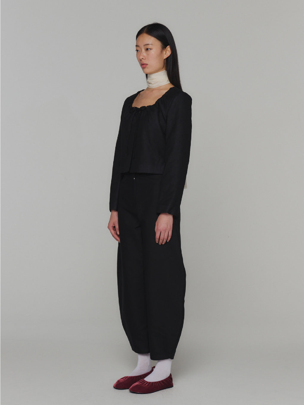 A "Sheer Padded Cropped Jacket Black" by Amomento displayed on a white background. The jacket, with its black color and long sleeves, boasts an adjustable square neckline, natural shirring near the chest area, and a subtle bow detail at the front. Its minimalistic design is both elegant and contemporary.