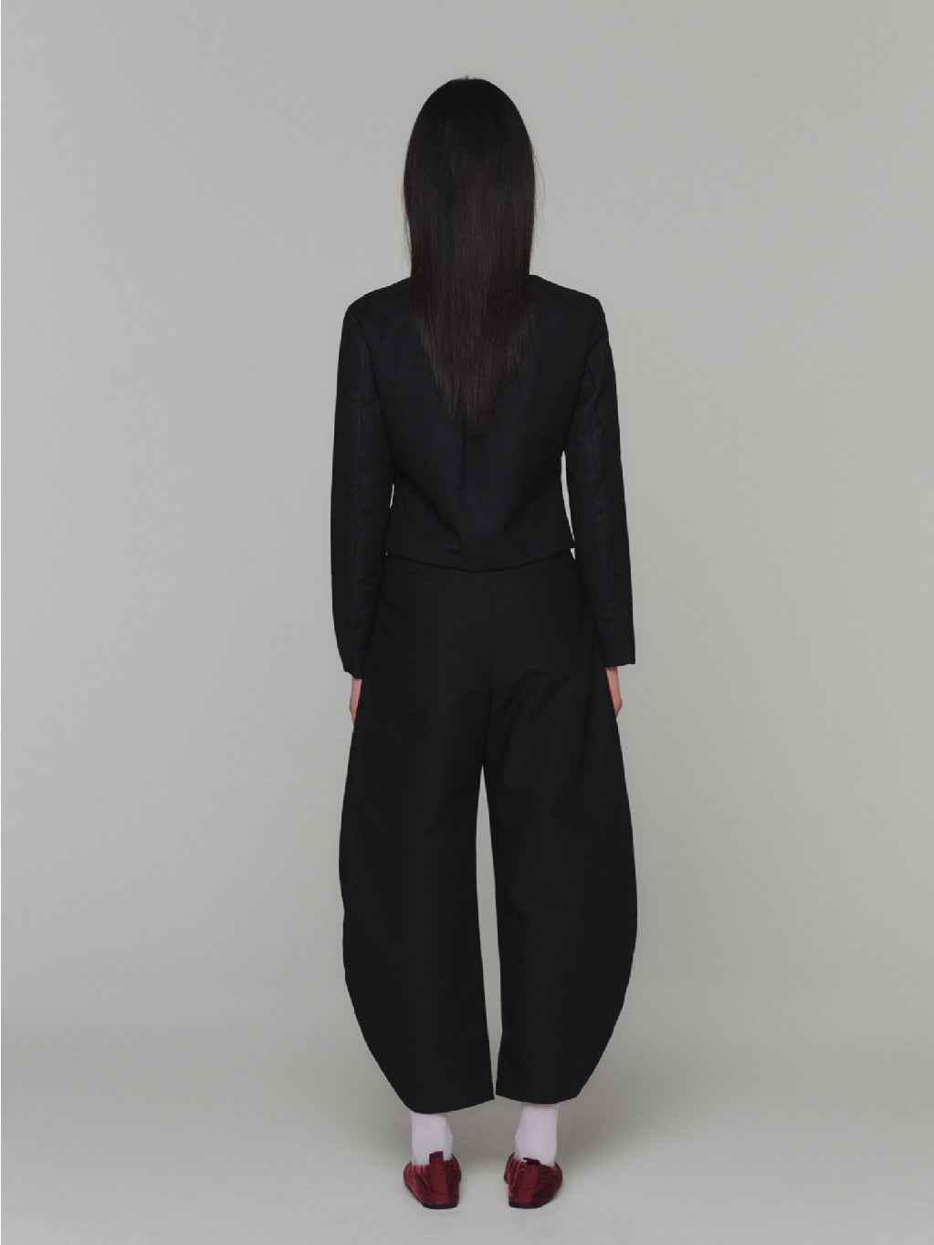 A "Sheer Padded Cropped Jacket Black" by Amomento displayed on a white background. The jacket, with its black color and long sleeves, boasts an adjustable square neckline, natural shirring near the chest area, and a subtle bow detail at the front. Its minimalistic design is both elegant and contemporary.