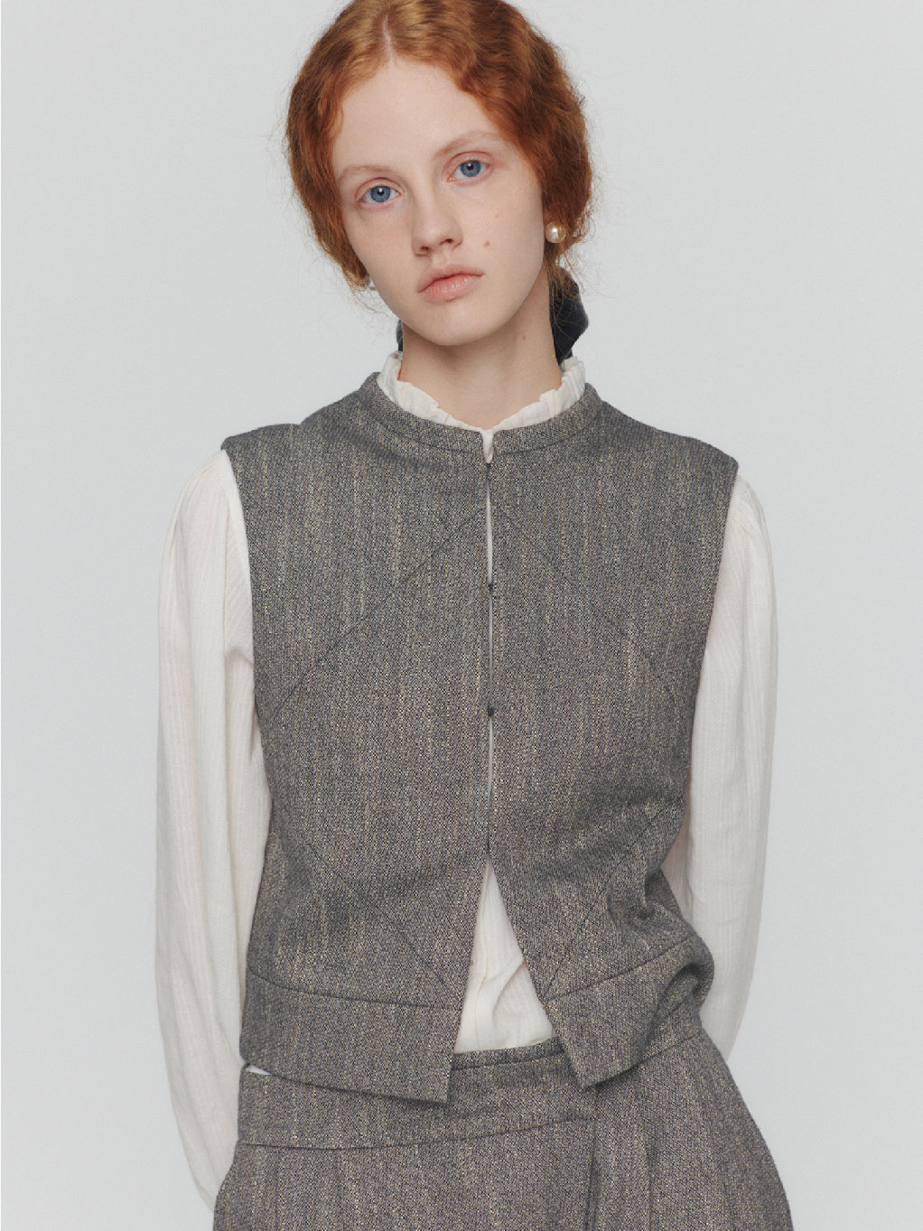 The Wool Tweed Motive Vest Grey by Amomento is a sleeveless, boxy, cropped vest made from a wool blend with a textured or woven appearance. It features subtle diamond-shaped stitch details and has a round neckline with no visible buttons or zippers on the front.