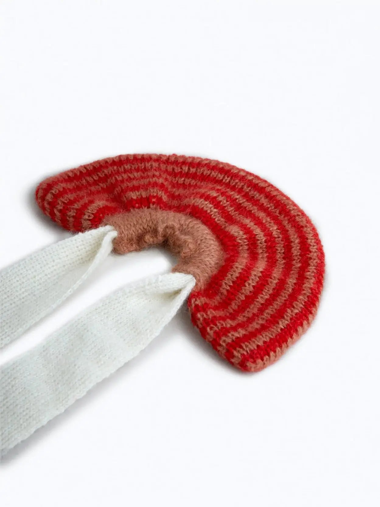 A whimsical Amapola Mohair Bow Scrunchie from Tomasa, colored in red and pink stripes, features long white knitted ties extending downwards. The scrunchie has a unique semi-circular shape, reminiscent of a rainbow. Available at our Barcelona store, the long ties are designed to wrap around for added warmth and style.