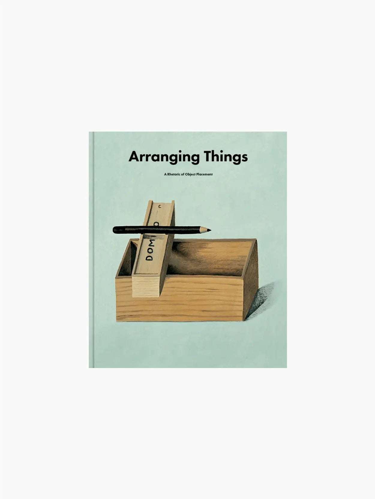 Book cover of "Arranging Things: A Rhetoric of Object Placement" by Apartamento. The cover features a wooden block partially inside a wooden box with additional blocks stacked above it. A pencil rests on top, and the background is a light pastel blue, reminiscent of a stylish Barcelona store display.