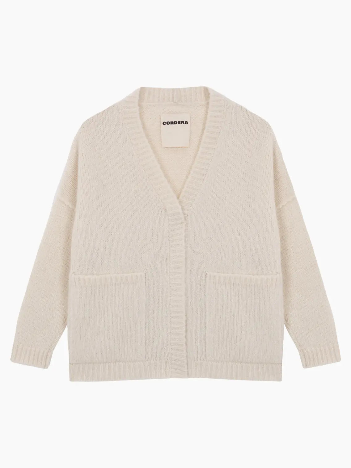 A cream-colored, long-sleeve Baby Alpaca Cardigan Natural displayed on a white background. It features a deep V-neck, front button closure, ribbed cuffs, two front pockets, and a relaxed fit. The label "Cordera" is visible on the inside neckline. Available exclusively at Bassalstore in Barcelona.