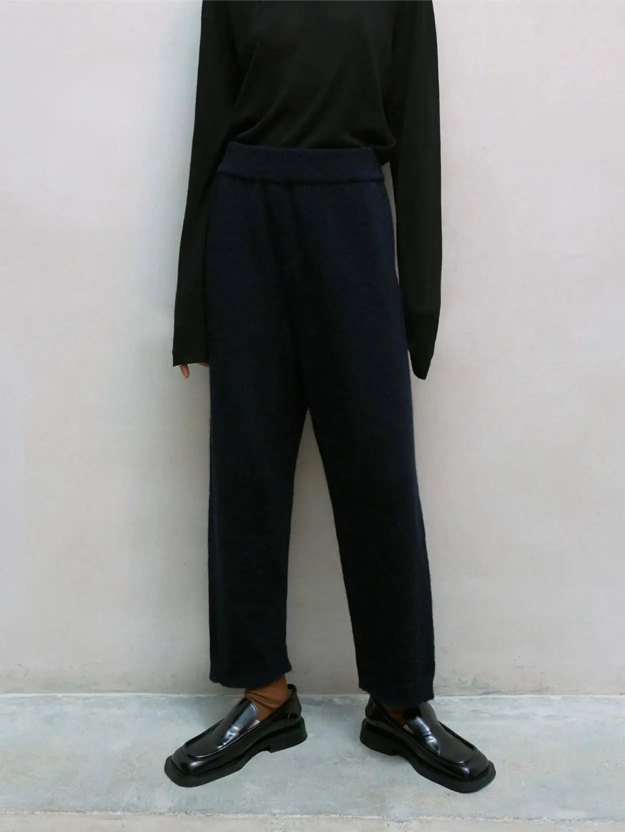 A person is standing against a white wall, wearing a black long-sleeve top, Baby Alpaca Knit Pants Navy from Cordera, and black shiny loafers. The person’s head is not shown in the image.