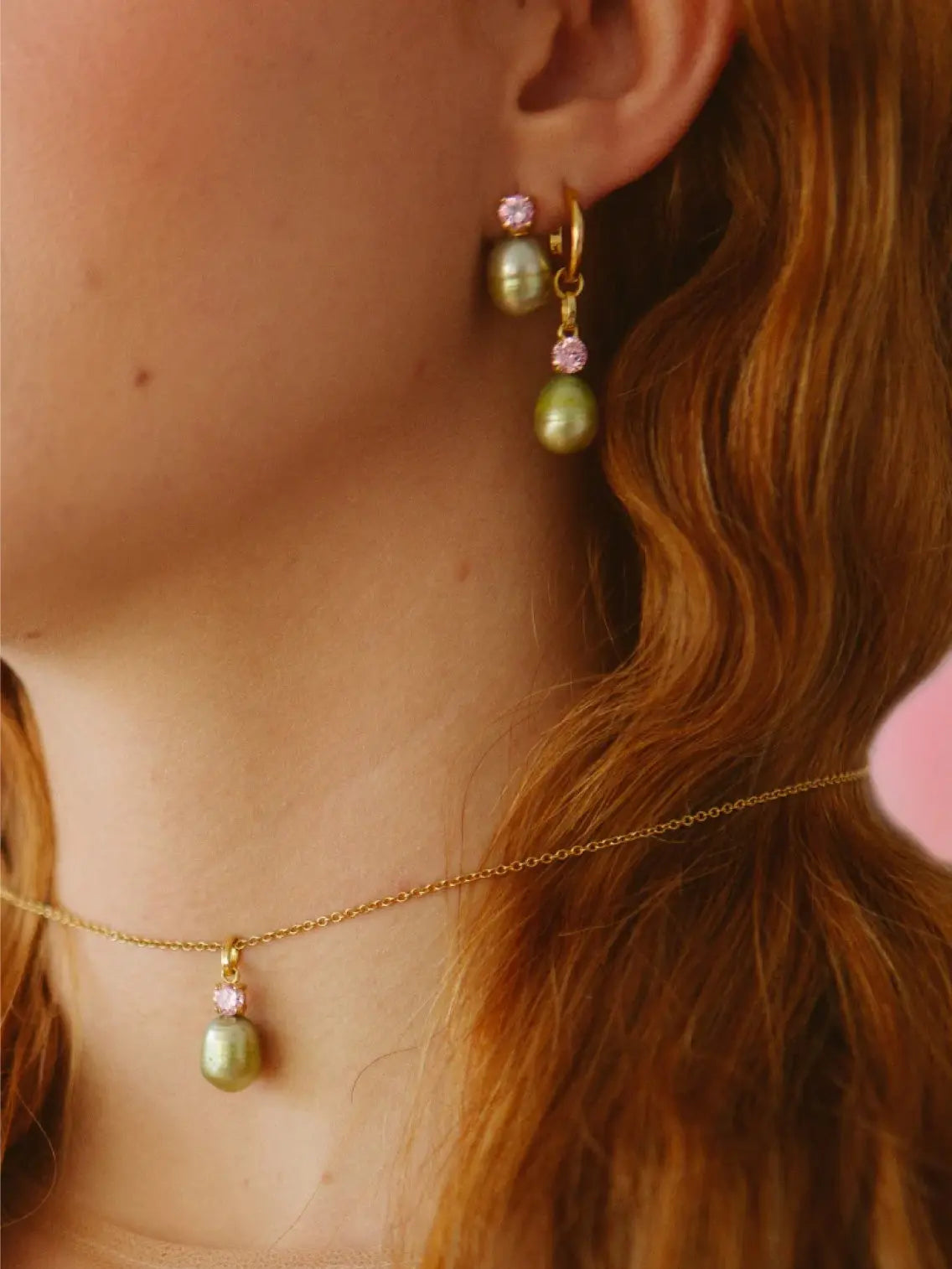A gold hoop earring from Wilhelmina Garcia, called the Bailarina Hoop Earring, features a hanging light green pearl and a small pink gemstone above it. The earring has an inscription "925" on the inner side of the hoop. The background is white.