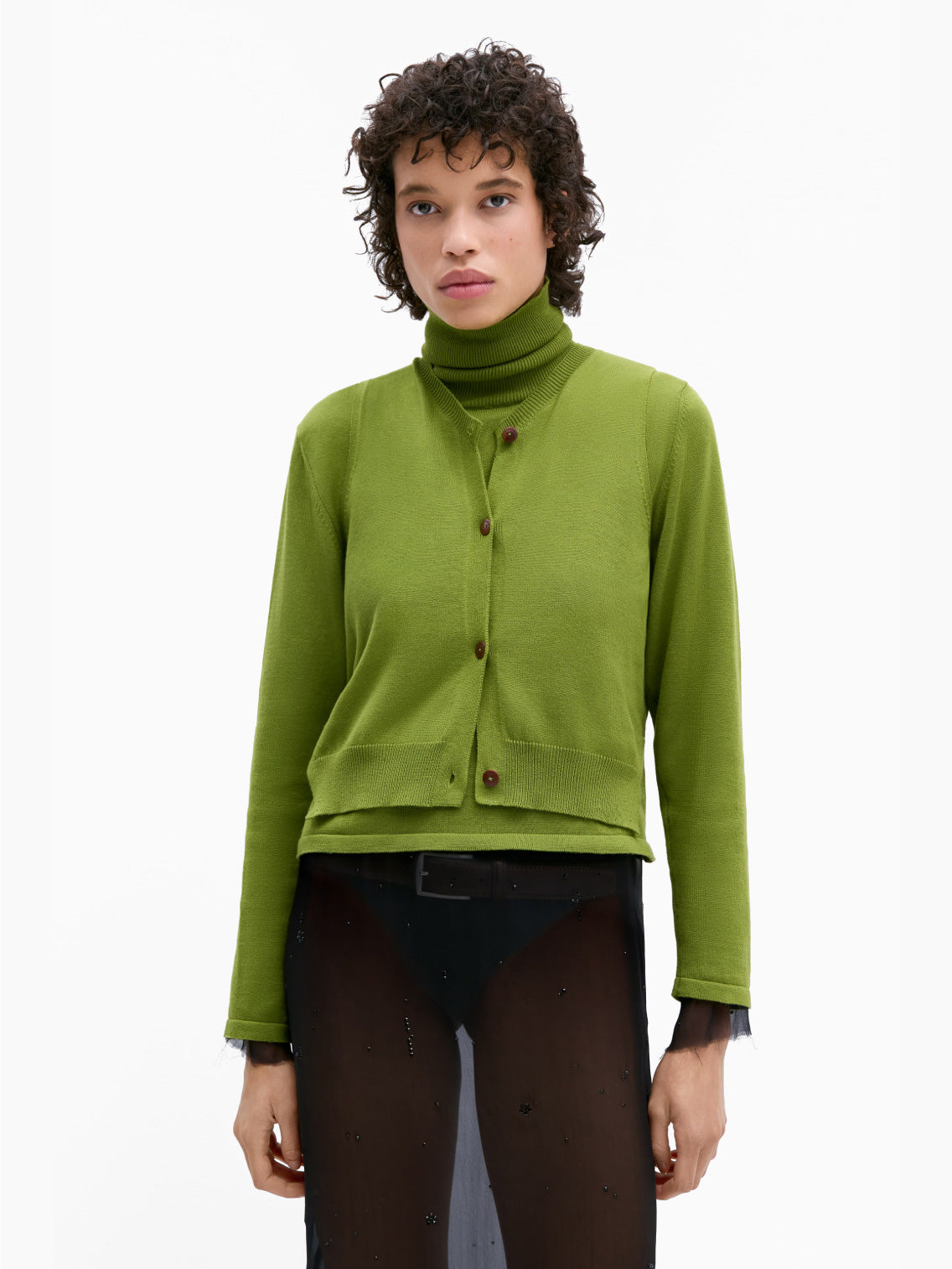 A person with short, curly hair is wearing Cordera's Cotton & Cashmere Waistcoat in Woodbine, a bright green V-neck sweater made from an opulent cashmere blend, buttoned up over a matching turtleneck. They pair this with a sheer black skirt featuring a black underlayer and are standing against a plain white background.