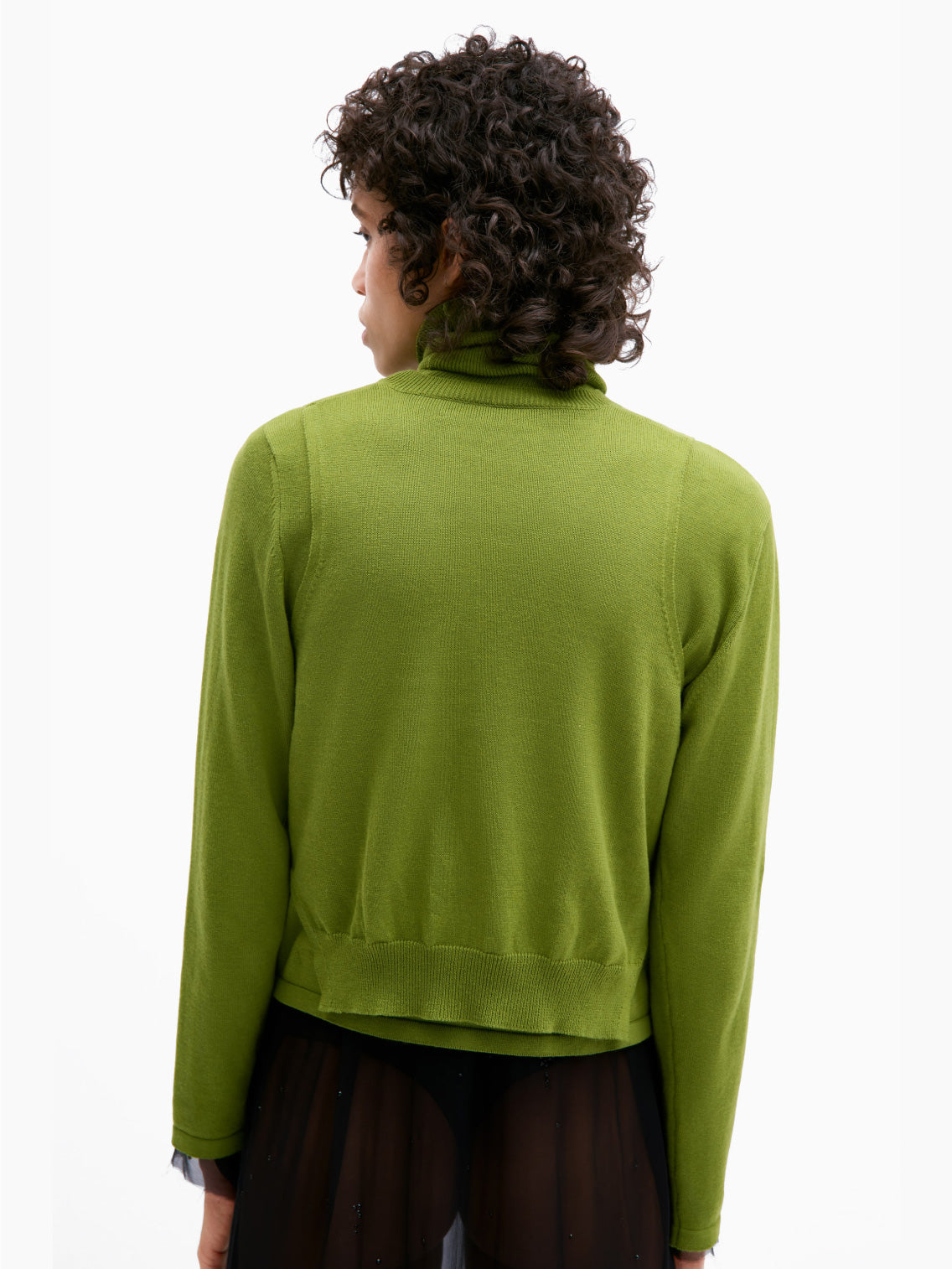 A person with short, curly hair is wearing Cordera's Cotton & Cashmere Waistcoat in Woodbine, a bright green V-neck sweater made from an opulent cashmere blend, buttoned up over a matching turtleneck. They pair this with a sheer black skirt featuring a black underlayer and are standing against a plain white background.