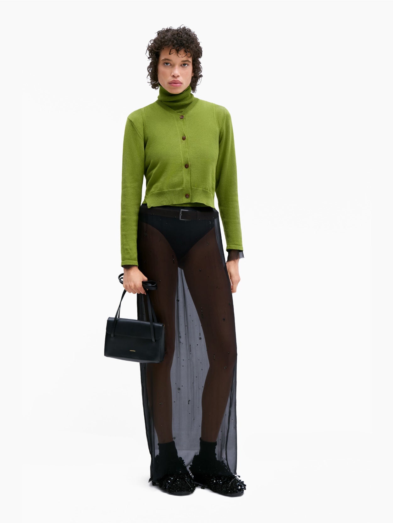 A person with short, curly hair is wearing Cordera's Cotton & Cashmere Waistcoat in Woodbine, a bright green V-neck sweater made from an opulent cashmere blend, buttoned up over a matching turtleneck. They pair this with a sheer black skirt featuring a black underlayer and are standing against a plain white background.