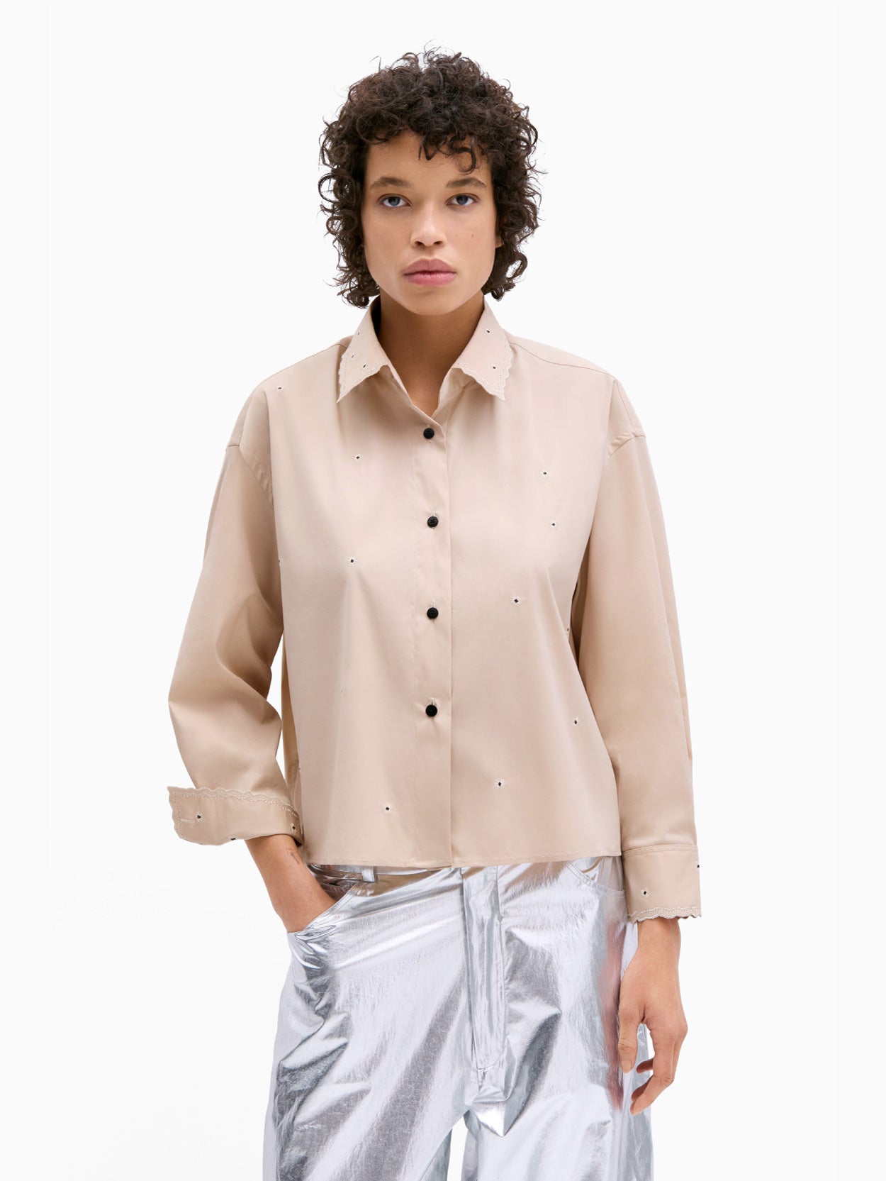 A person with curly hair is wearing the Cordera Embroidery Shirt Beige, an oversized fit shirt featuring small holes and intricate metallic thread embroidery. They have paired it with silver pants and are standing against a plain white background, one hand in their pocket, looking directly at the camera.