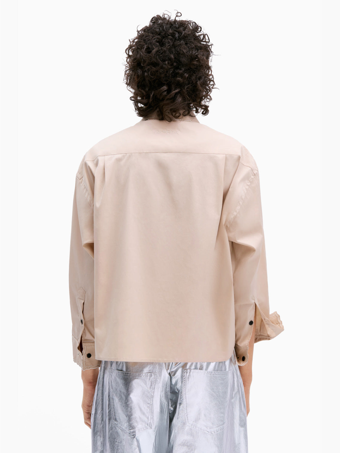 A person with curly hair is wearing the Cordera Embroidery Shirt Beige, an oversized fit shirt featuring small holes and intricate metallic thread embroidery. They have paired it with silver pants and are standing against a plain white background, one hand in their pocket, looking directly at the camera.