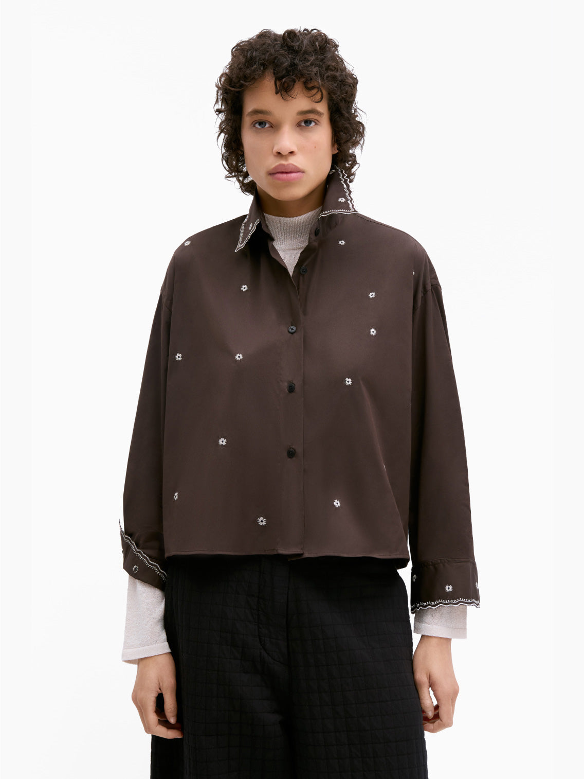 A person with short, curly hair is wearing the "Embroidery Shirt Chocolate" by Cordera over a light-colored turtleneck. The dark brown, long-sleeve shirt features white floral patterns and metallic thread embroidery. Its cropped and oversized fit adds a unique touch as they stand against a plain white background.