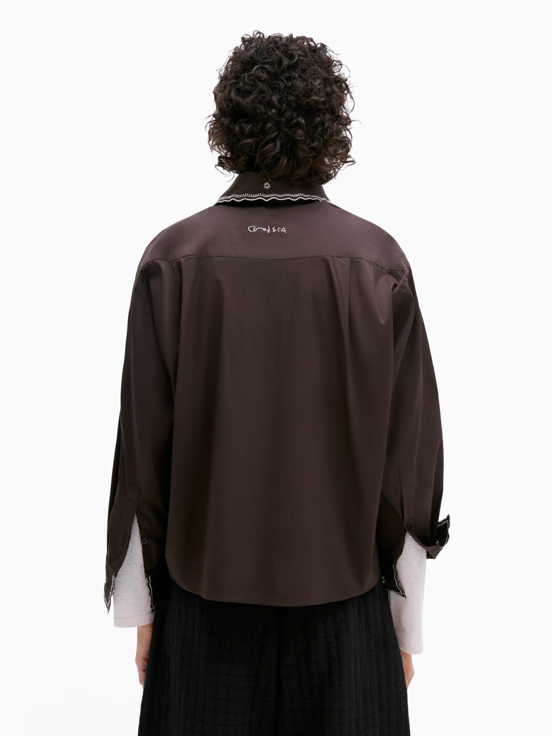 A person with short, curly hair is wearing the "Embroidery Shirt Chocolate" by Cordera over a light-colored turtleneck. The dark brown, long-sleeve shirt features white floral patterns and metallic thread embroidery. Its cropped and oversized fit adds a unique touch as they stand against a plain white background.