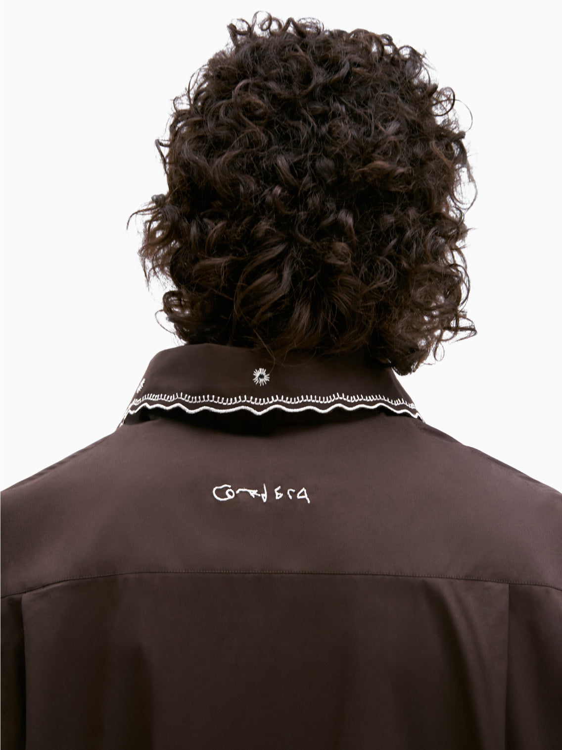 A person with short, curly hair is wearing the "Embroidery Shirt Chocolate" by Cordera over a light-colored turtleneck. The dark brown, long-sleeve shirt features white floral patterns and metallic thread embroidery. Its cropped and oversized fit adds a unique touch as they stand against a plain white background.