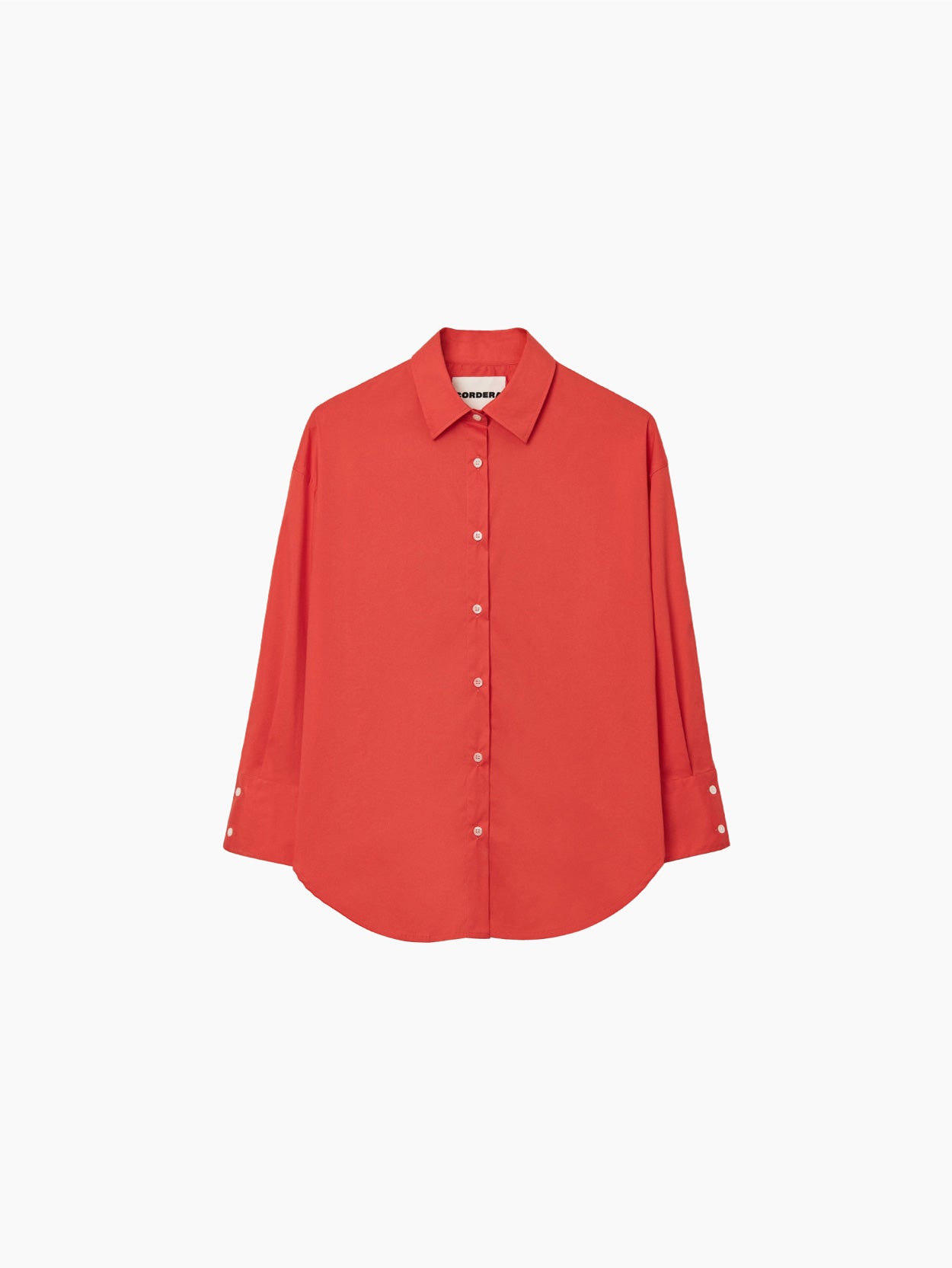 The Cordera Oversize Cotton Shirt in coral is a long-sleeved, pure cotton garment. It features a pointed collar and cuffed sleeves with buttons. Crafted from lightweight fabric, it has an oversized silhouette and a curved hemline. The small, white buttons provide a striking contrast against the coral fabric.