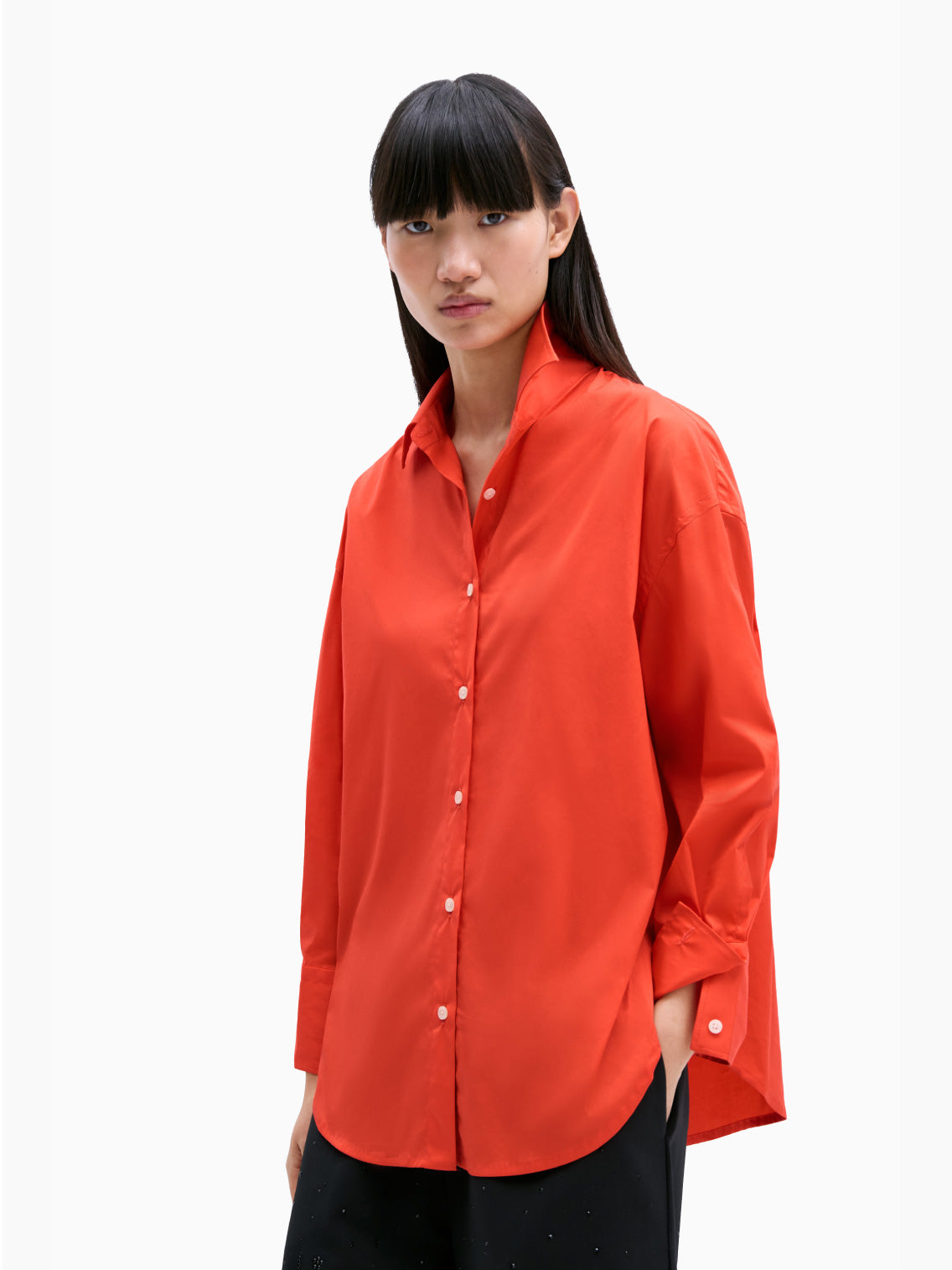 The Cordera Oversize Cotton Shirt in coral is a long-sleeved, pure cotton garment. It features a pointed collar and cuffed sleeves with buttons. Crafted from lightweight fabric, it has an oversized silhouette and a curved hemline. The small, white buttons provide a striking contrast against the coral fabric.