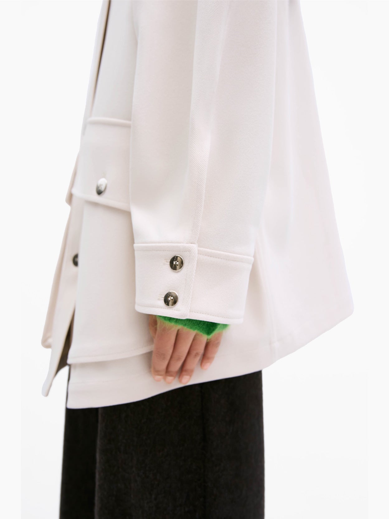 A person with long, dark hair stands facing forward against a plain white background. They are wearing an oversize fit Short Trench Coat Ecru by Cordera, featuring a brown collar, a green shirt underneath, loose dark pants, and white sneakers. Their hands are relaxed by their sides.