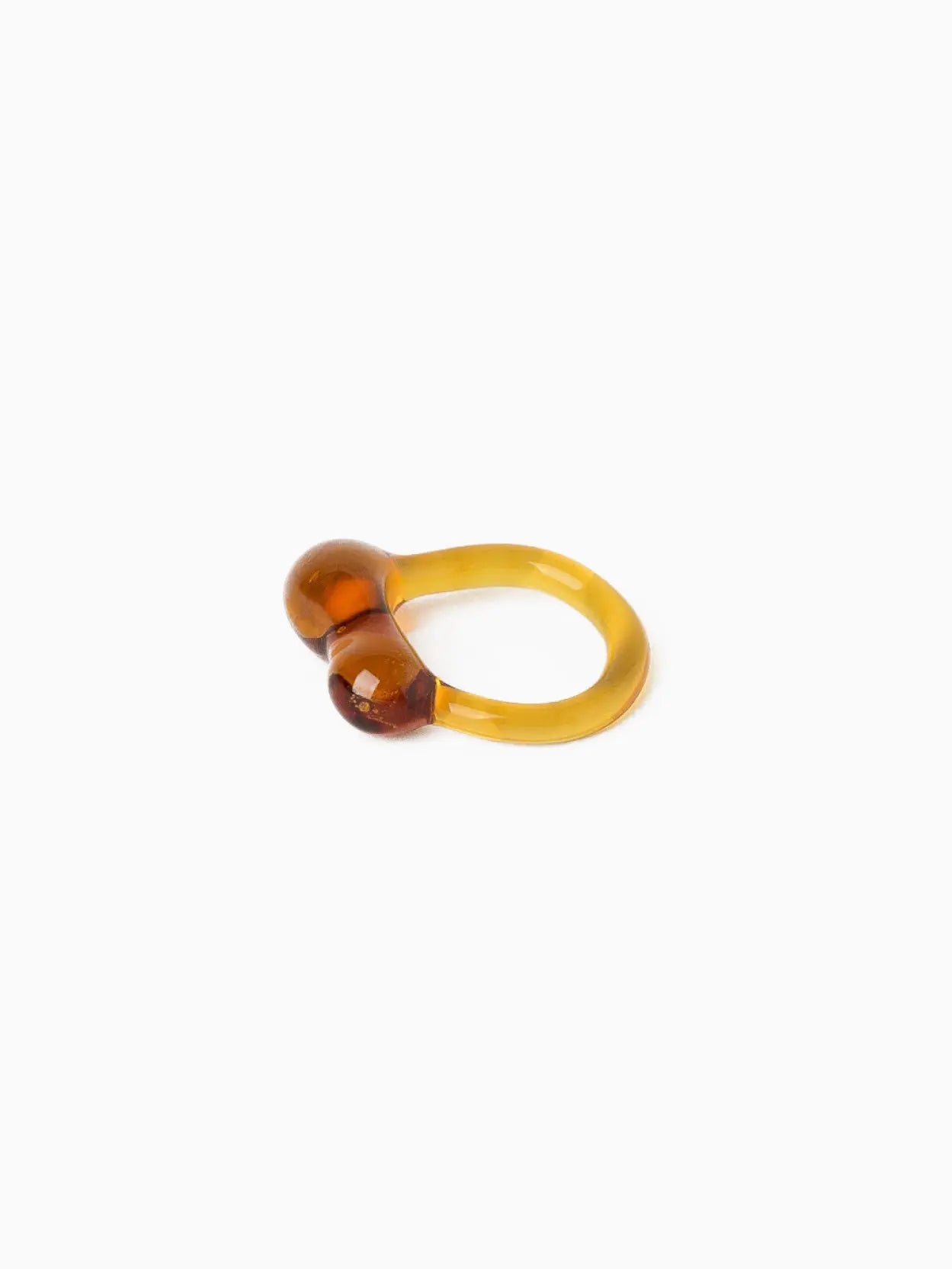 A minimalist, amber-colored Caju Ring Amber made of translucent resin by Nathalie Schreckenberg available at Bassalstore. The ring features two bulbous ends positioned opposite each other on the band. The simple yet elegant design gives the ring a modern and unique appearance. The background is plain white, reflecting its chic Barcelona roots.