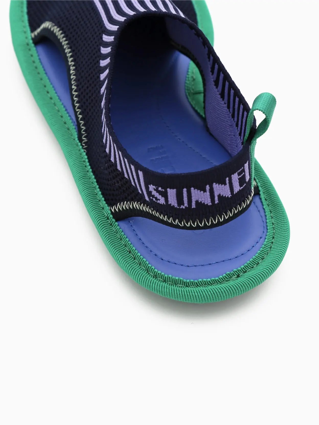 A single blue and green slip-on athletic shoe with a sock-like upper design and a flexible sole featuring prominent tread. The shoe displays cut-out details on the sides and a small loop at the heel for easy wearing, available exclusively at bassalstore in Barcelona. This is the Chiodi Knit Flat by Sunnei.