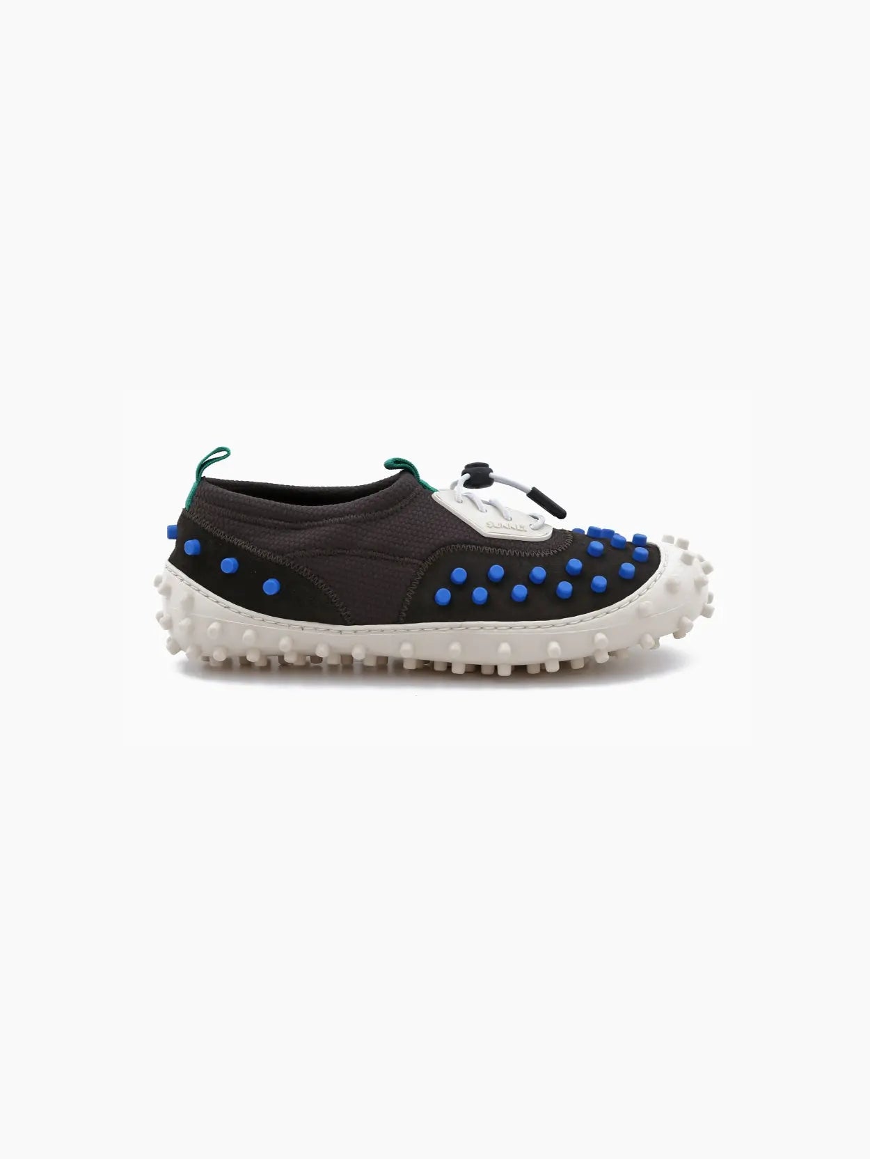 Side view of a unique sneaker from bassalstore featuring a black upper with blue and white patches, and green accents near the heel. The shoe has a white rubber sole adorned with large protruding nubs, creating a textured, rugged appearance. Perfect for your next visit to Barcelona. This is the Sunnei 1000 Chiodi TRK.