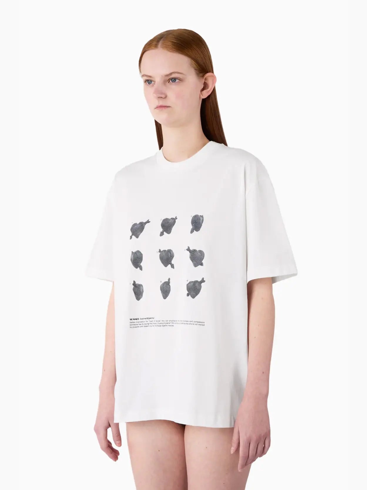 A white short-sleeved Classic T-Shirt "Cuori di Pietra" by Sunnei with a minimalist design featuring nine black abstract shapes arranged in a grid pattern on the front. There is text below the shapes. Available exclusively at Bassalstore, your go-to store in Barcelona.