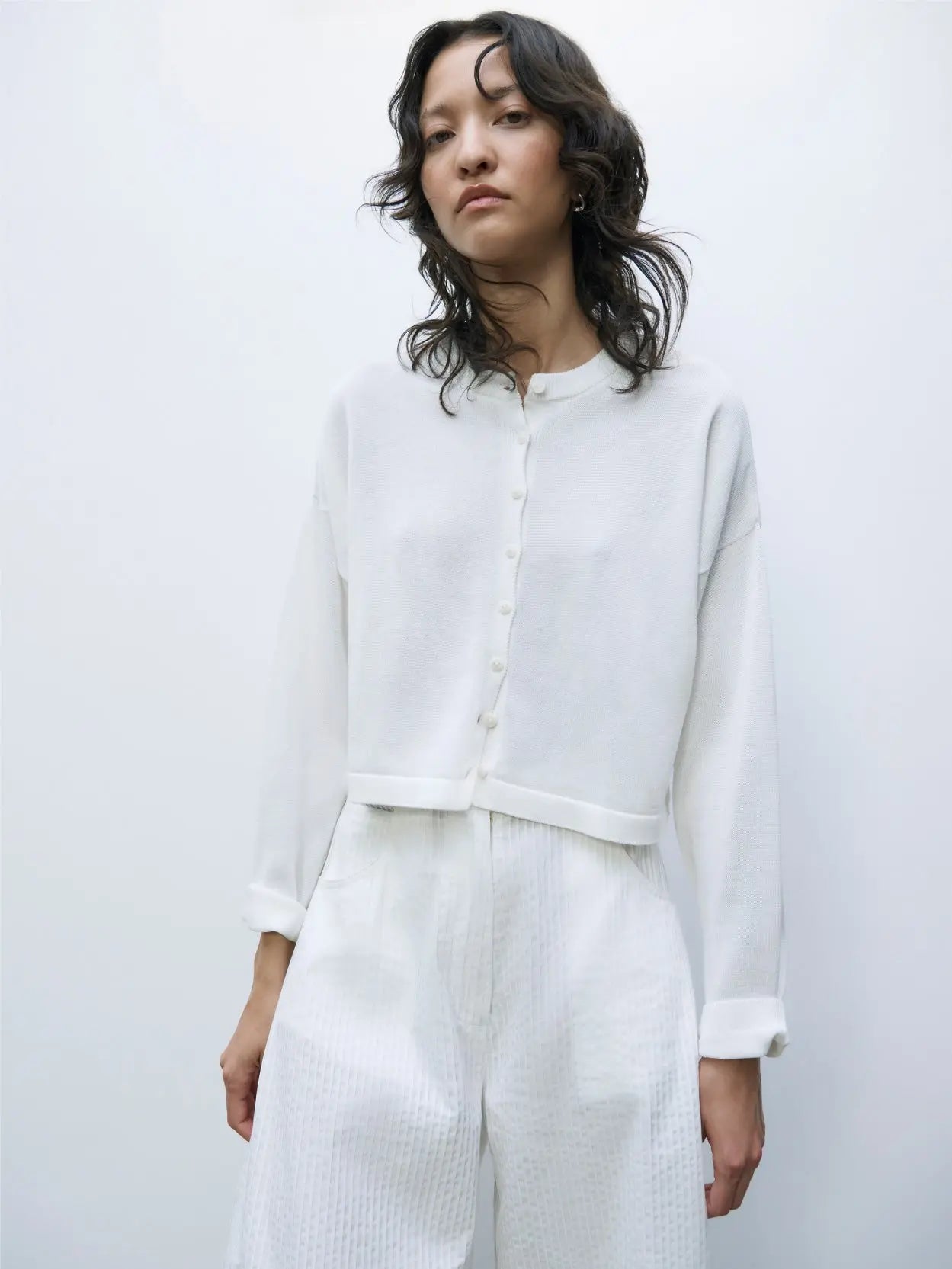 A white, long-sleeved, cropped cardigan with buttons down the front from Cordera. The Cotton Cropped Cardigan White has a simple, minimalist design with a round neckline and ribbed cuffs at the sleeves. The fabric appears soft and lightweight against a plain white background.