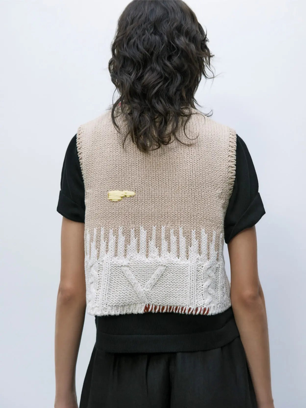 A beige knitted sleeveless vest with a textured pattern, perfect for adding a touch of Barcelona flair to your wardrobe. The upper part is plain, while the lower part features white knitted designs resembling drips. Small, colorful abstract shapes are scattered on the vest, available exclusively at Bassalstore as the Cotton Embroidered Top by Cordera.