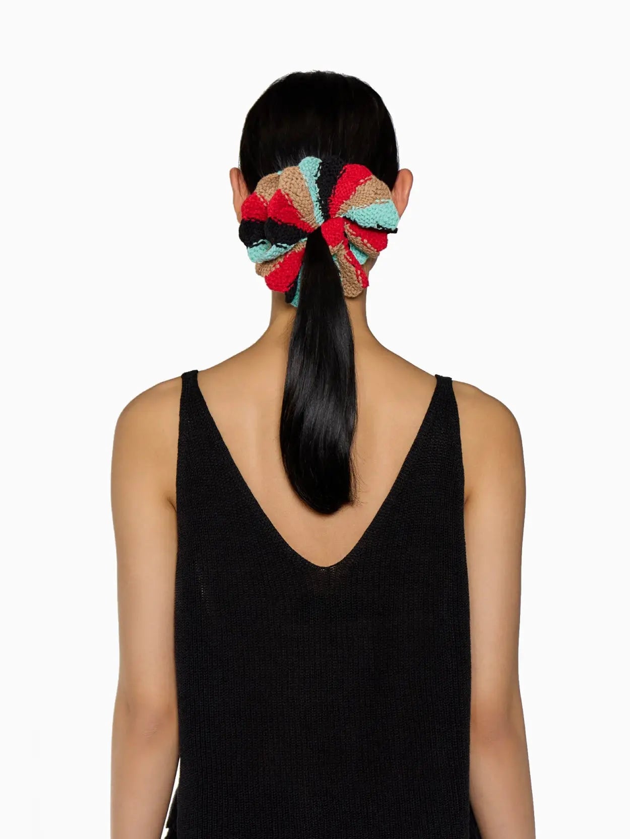 A multicolored, crocheted Fernandita Scrunchie Berber made up of alternating segments in red, black, blue, and beige. It is circular and has a visible fabric brand tag labeled "Tomasa." Available at bassalstore in Barcelona. The background is plain white.