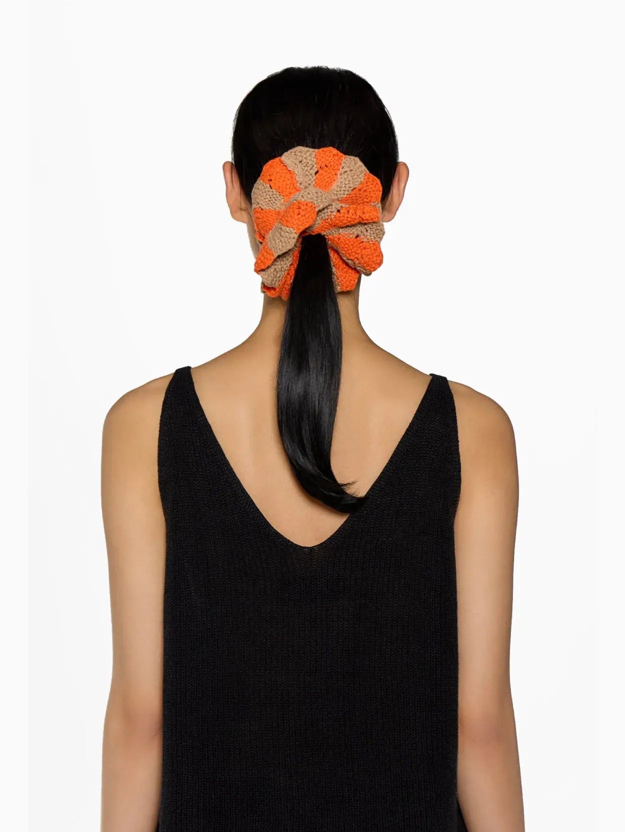 A round, knitted scrunchie in alternating orange and beige segments with a small white label that reads "Tomasa." The Fernandita Scrunchie, available at Bassalstore Barcelona, is placed vertically in the center of a white background.