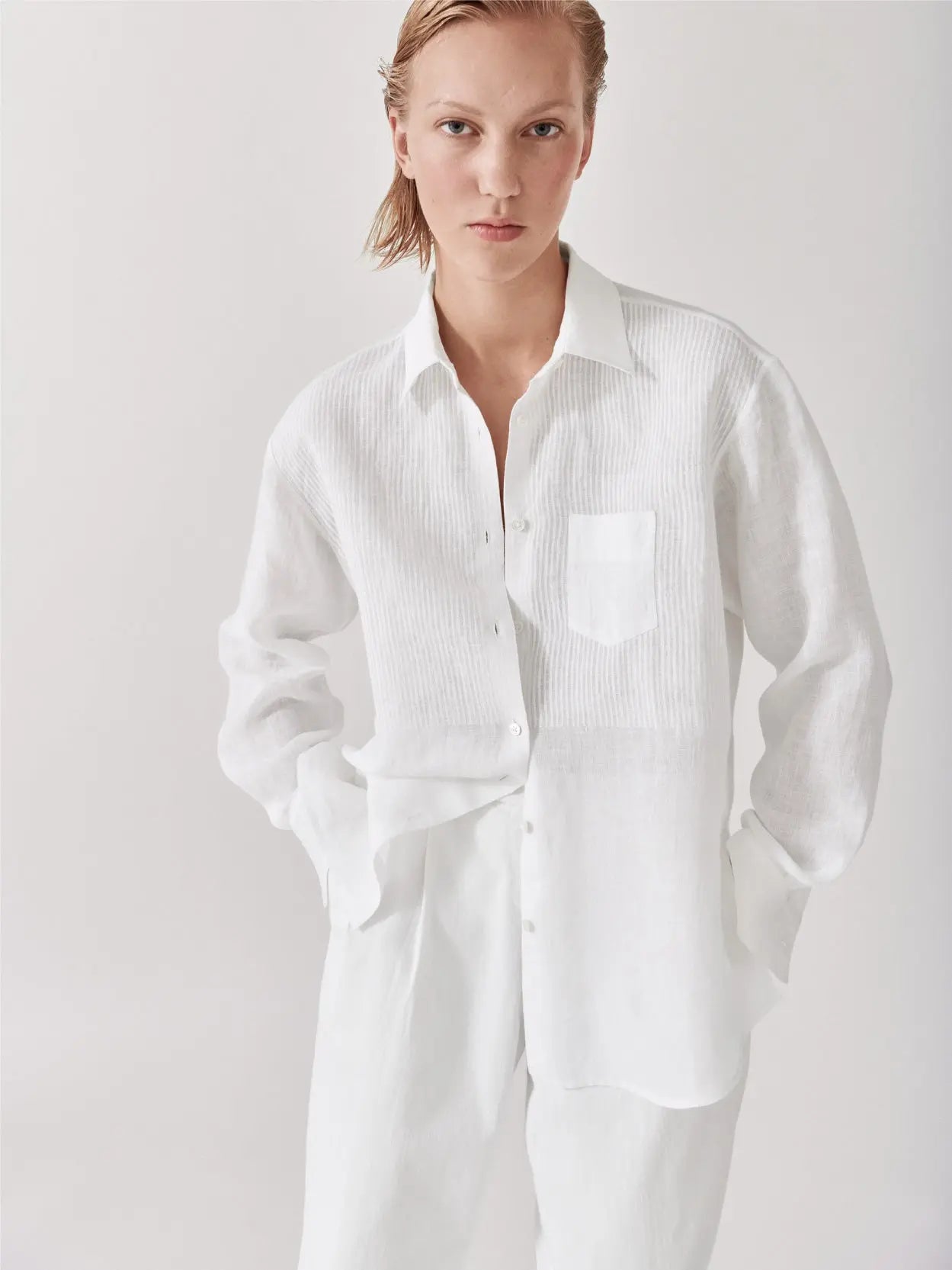 A white long-sleeve button-up shirt with a single chest pocket and a classic collar is neatly displayed against a plain white background. The Galla Linen Shirt Block Print Stripe by Jan Machenhauer, available at Bassalstore in Barcelona, features a relaxed fit with slightly rounded hems.