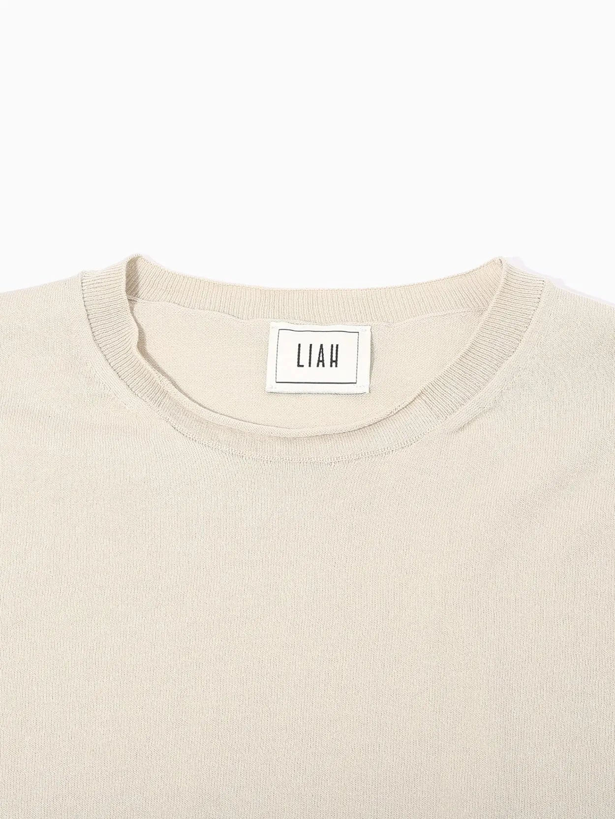 A beige, long-sleeved sweater from Bassalstore is displayed against a plain white background. The Giada T-Shirt Tea from Liah has a minimalist design, featuring a round neckline, ribbed cuffs, and a slightly loose fit. There's a white label with branding at the back of the neckline, reflecting its Barcelona origins.