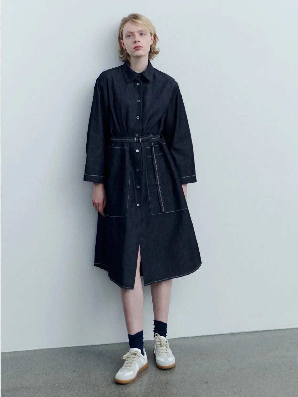 The Giselle Denim Dress by Jan Machenhauer is a dark denim, button-down shirt dress with long sleeves, a collar, and a waist belt. The dress features two front pockets, white buttons down the front, and a flared skirt. Available at BassalStore in Barcelona.