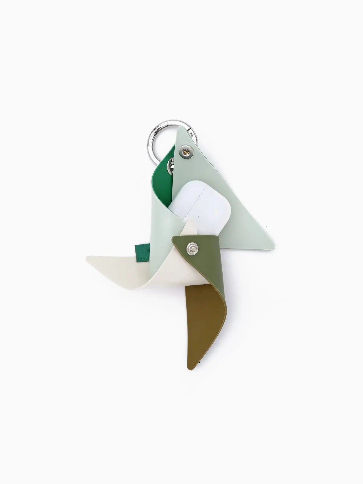 A minimalist, abstract sculpture from Barcelona, made from four folded, colored metal sheets in green, white, and brown attached to a circular metallic ring. It creates a pinwheel-like appearance and is set against a plain white background. The Sunnei Gomma Girandola Airpods Holder Multi Grass Green is available exclusively at Bassalstore.