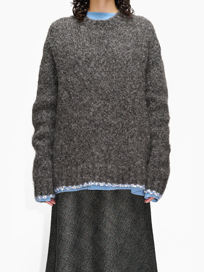 The Hope Box Sweater Grey, made from luxurious alpaca yarn, showcases a charcoal gray hue with subtle blue edging at the hem and cuffs. This knitted crewneck features a ribbed texture and classic design, laid flat against a white background.