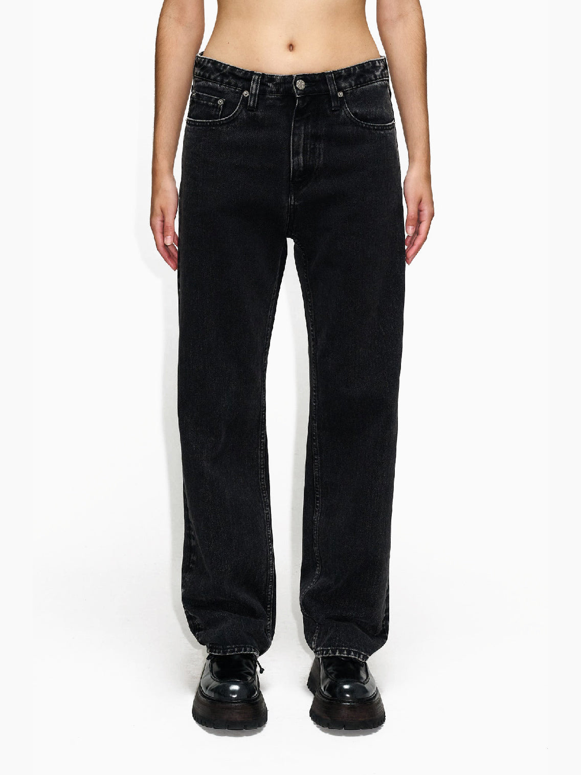 A person wearing Hope's Easy Jeans Chipped Black, high-waisted and loose-fitting, made from sustainable denim and paired with black shoes, stands with arms relaxed by their sides against a white background. These jeans, crafted from organic cotton, feature front pockets, belt loops, and a button/zipper closure. The person's torso is not shown.