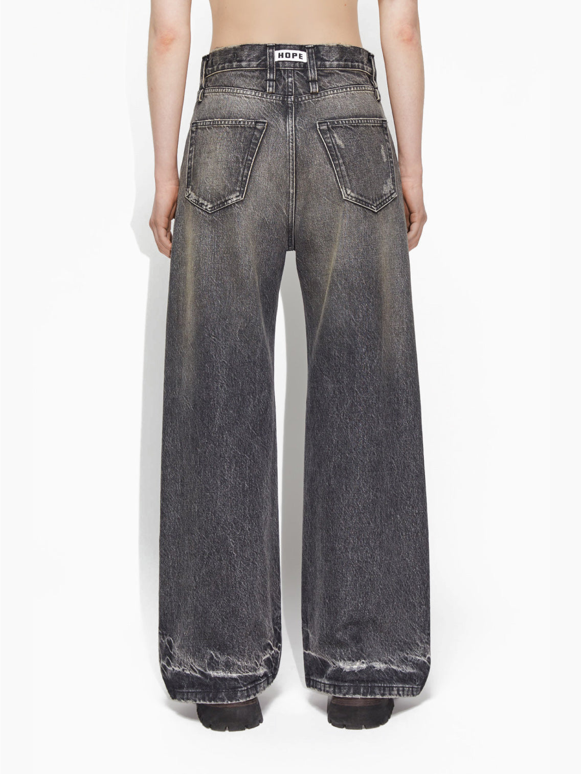 A person wearing Hope's Skid Jeans Heavy Black Vintage, which are loose-fitting and high-waisted with a slightly frayed hem. They are also sporting black shoes, while their upper body and head remain out of the frame against a plain white background.