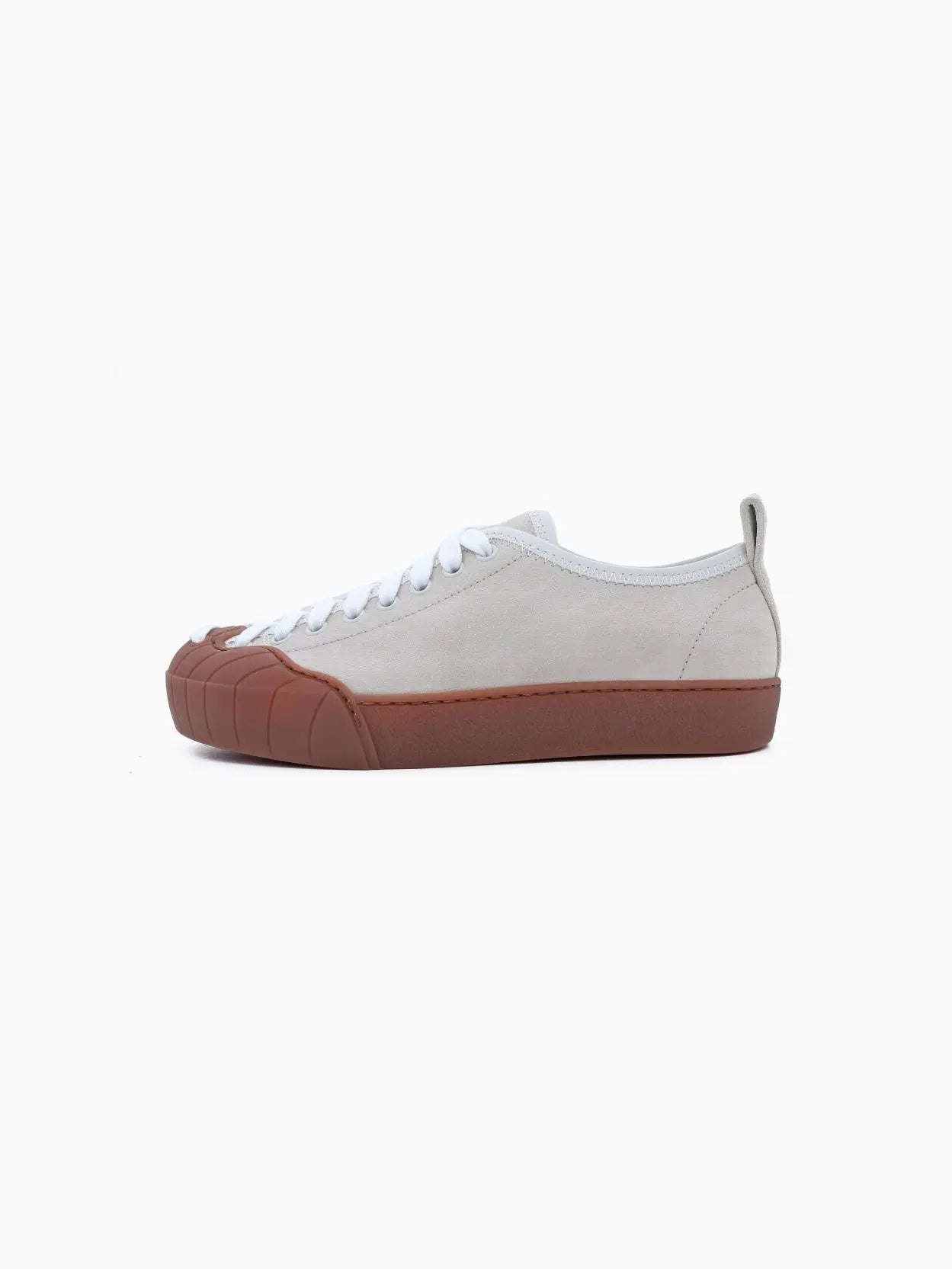 A side view of a single low-top sneaker with a beige upper, white laces, and a brown rubber sole that extends to cover the toe. The minimalist design includes a pull tab on the back for easy wear. Discover this chic **Sunnei Isi Low Off White** footwear at our Barcelona store or online at Bassalstore. The background is white.
