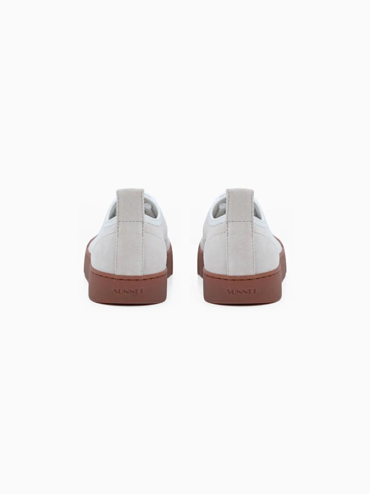 A side view of a single low-top sneaker with a beige upper, white laces, and a brown rubber sole that extends to cover the toe. The minimalist design includes a pull tab on the back for easy wear. Discover this chic **Sunnei Isi Low Off White** footwear at our Barcelona store or online at Bassalstore. The background is white.