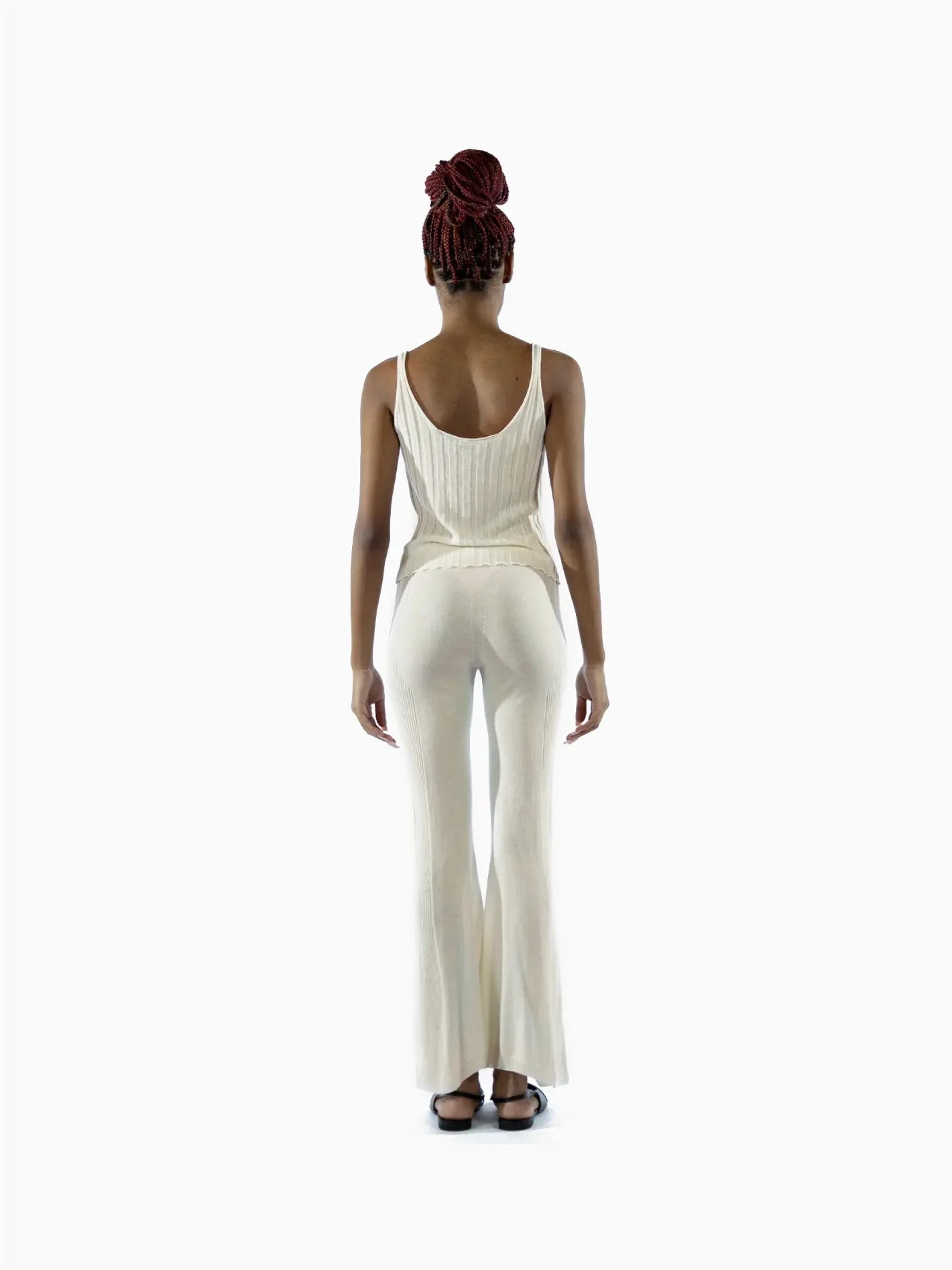 A person stands facing forward, dressed in a sleeveless, ribbed, cream-colored top and matching Lec Pants Ecru by Bielo with a slight flare. They have their hands by their sides and are wearing black shoes. The minimalist elegance of the outfit evokes the chic style reminiscent of Barcelona's fashion scene.