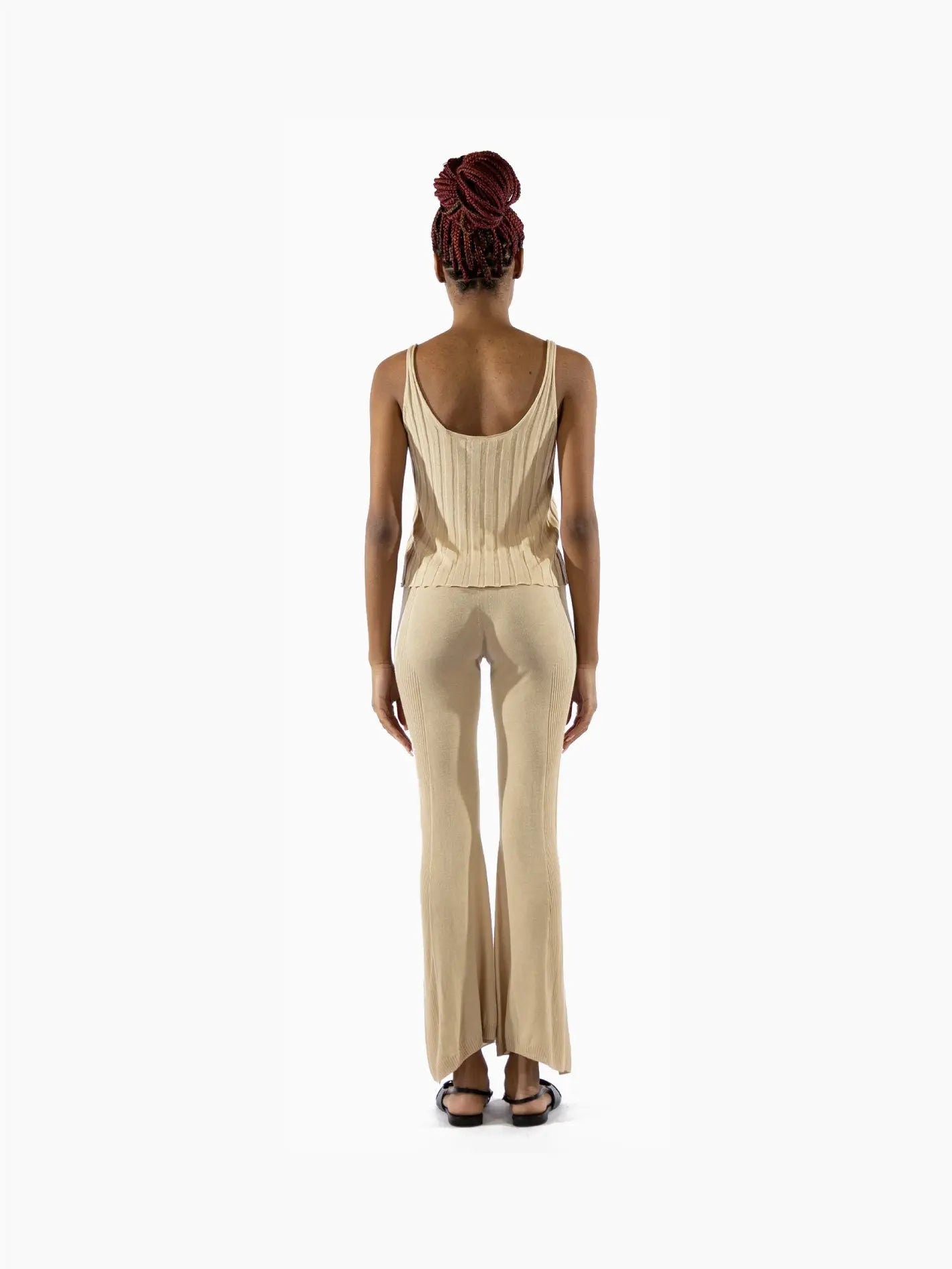 A person with an upright posture is wearing a beige knit tank top and matching wide-leg Lec Pants Latte by Bielo. They have dark hair styled in an updo and are standing against a plain white background, embodying the chic simplicity often seen in Barcelona's fashion. The outfit appears to be simple and comfortable.